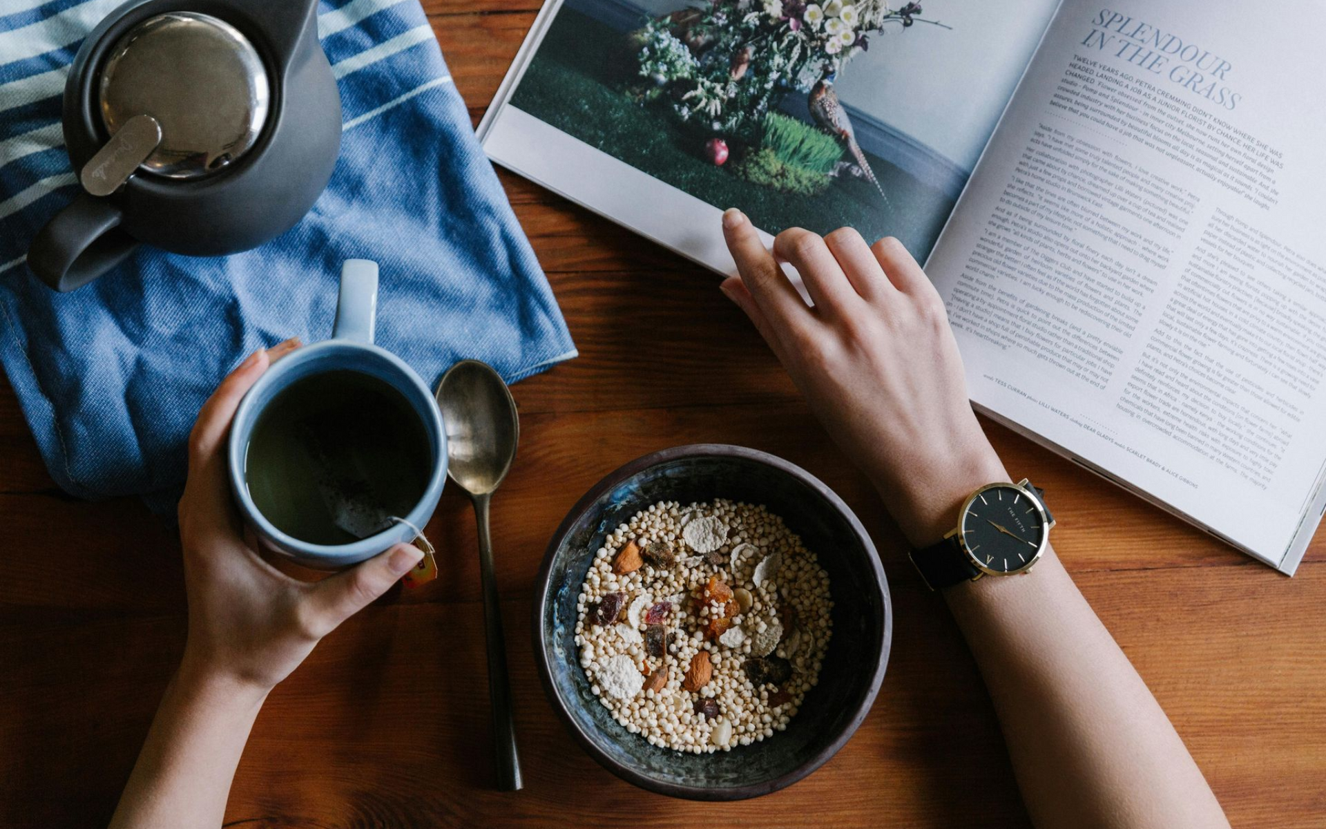 Person holding a mug, next to a bowl of cereal and a magazine on a wooden table.
