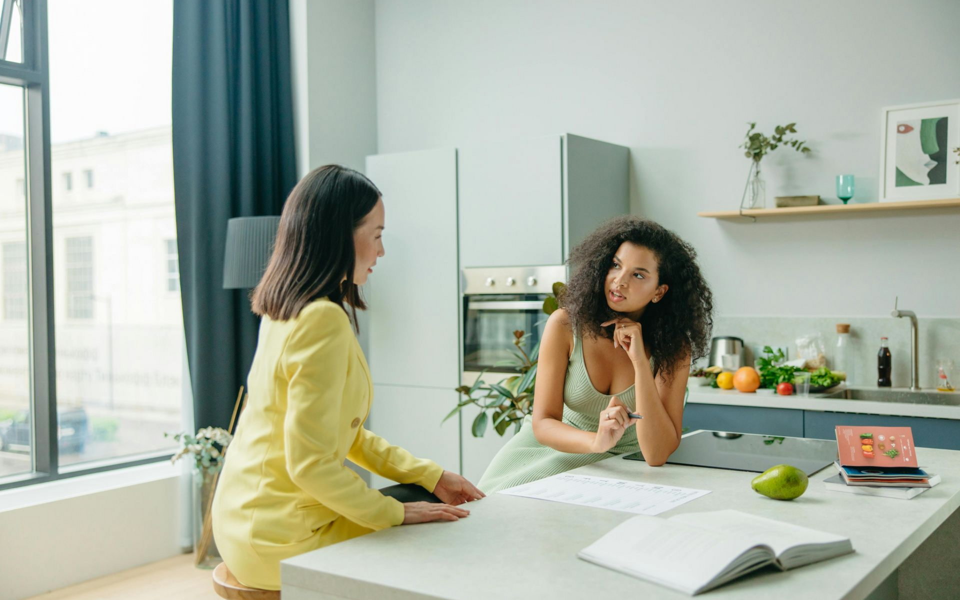 Two women in a kitchen, one in yellow jacket and one in a light dress, are talking at a counter with papers.