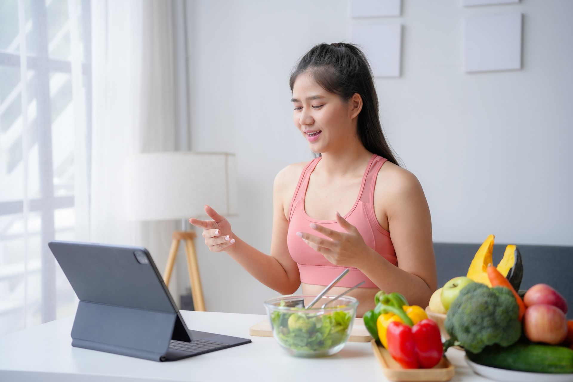 Woman in workout clothes gives online cooking lesson, surrounded by fresh produce.