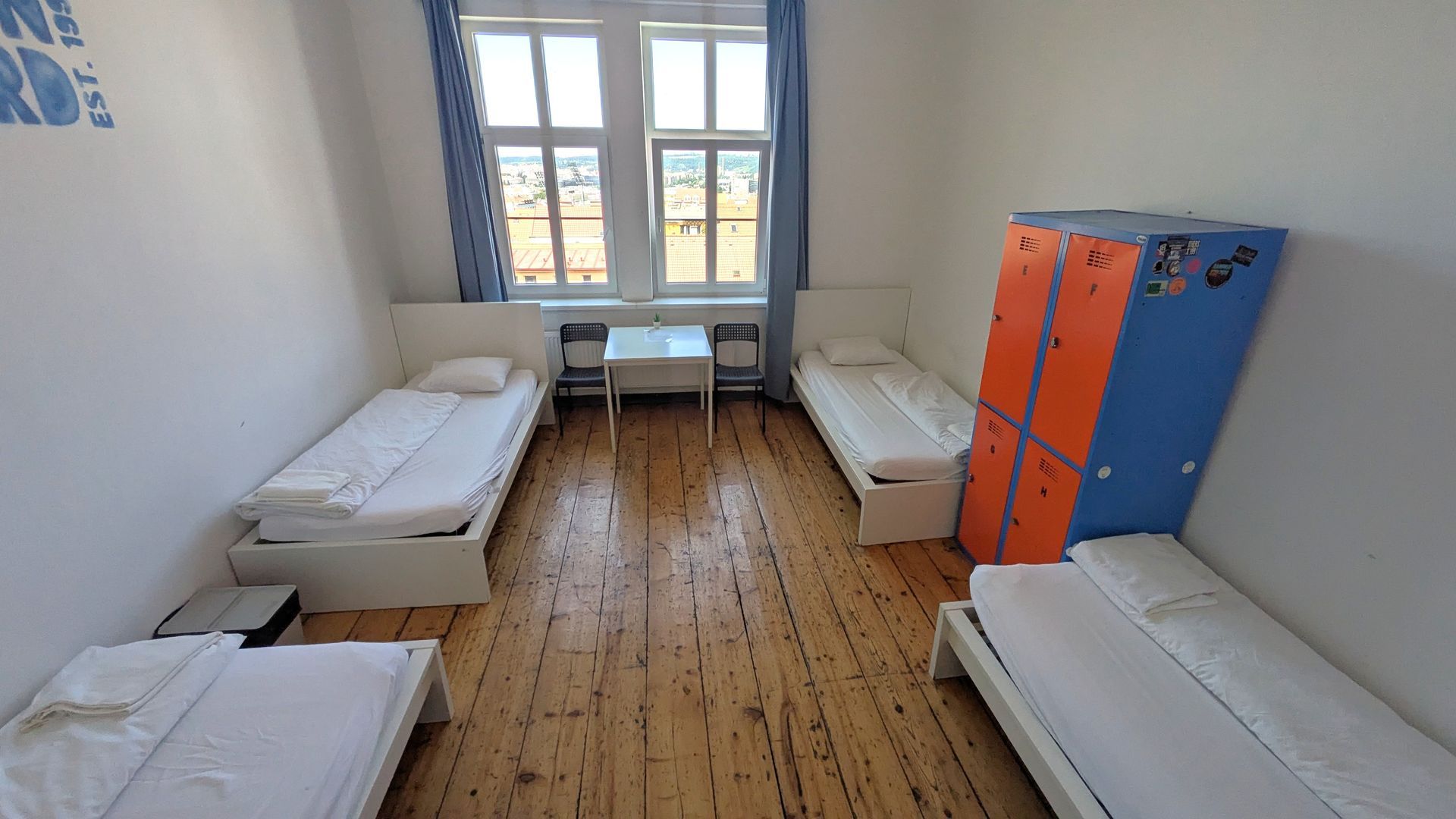 A hostel room with four beds, a window, and blue/orange lockers.