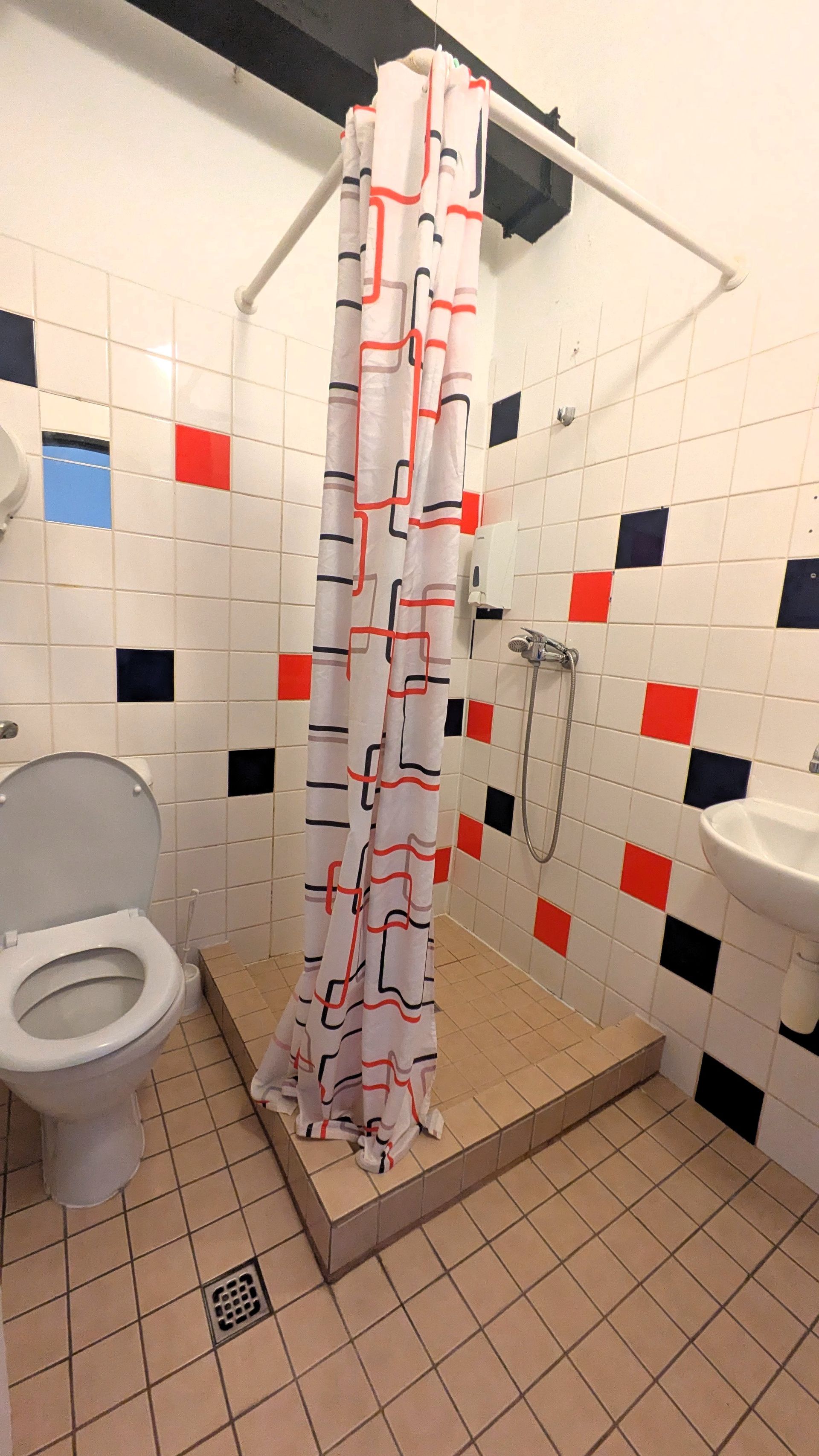 Bathroom with shower stall, toilet, sink, and tiles with red and blue squares. Shower curtain is patterned.