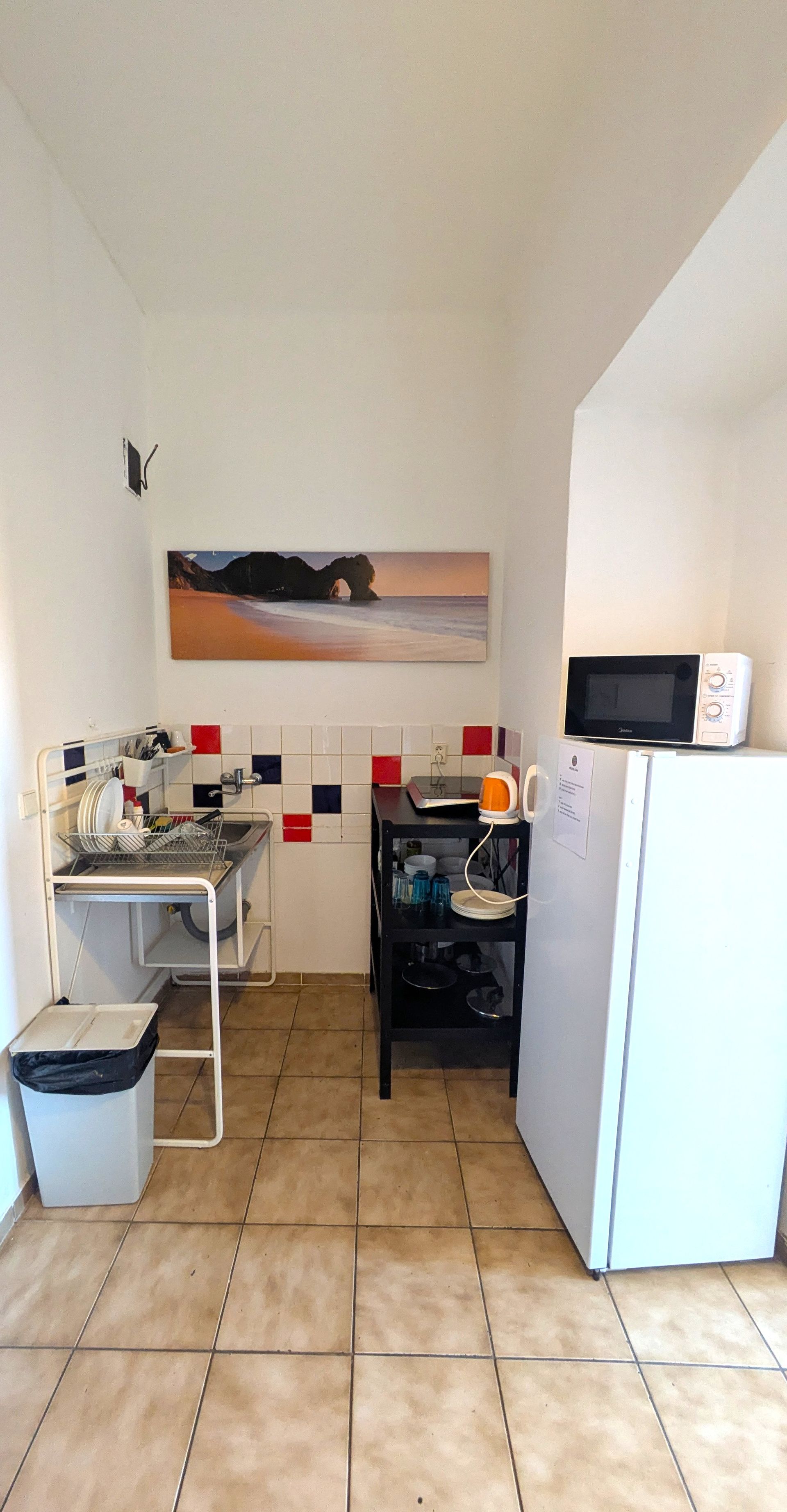 Small kitchen with a sink, microwave, refrigerator, and black shelving unit. Tile floor and white walls.