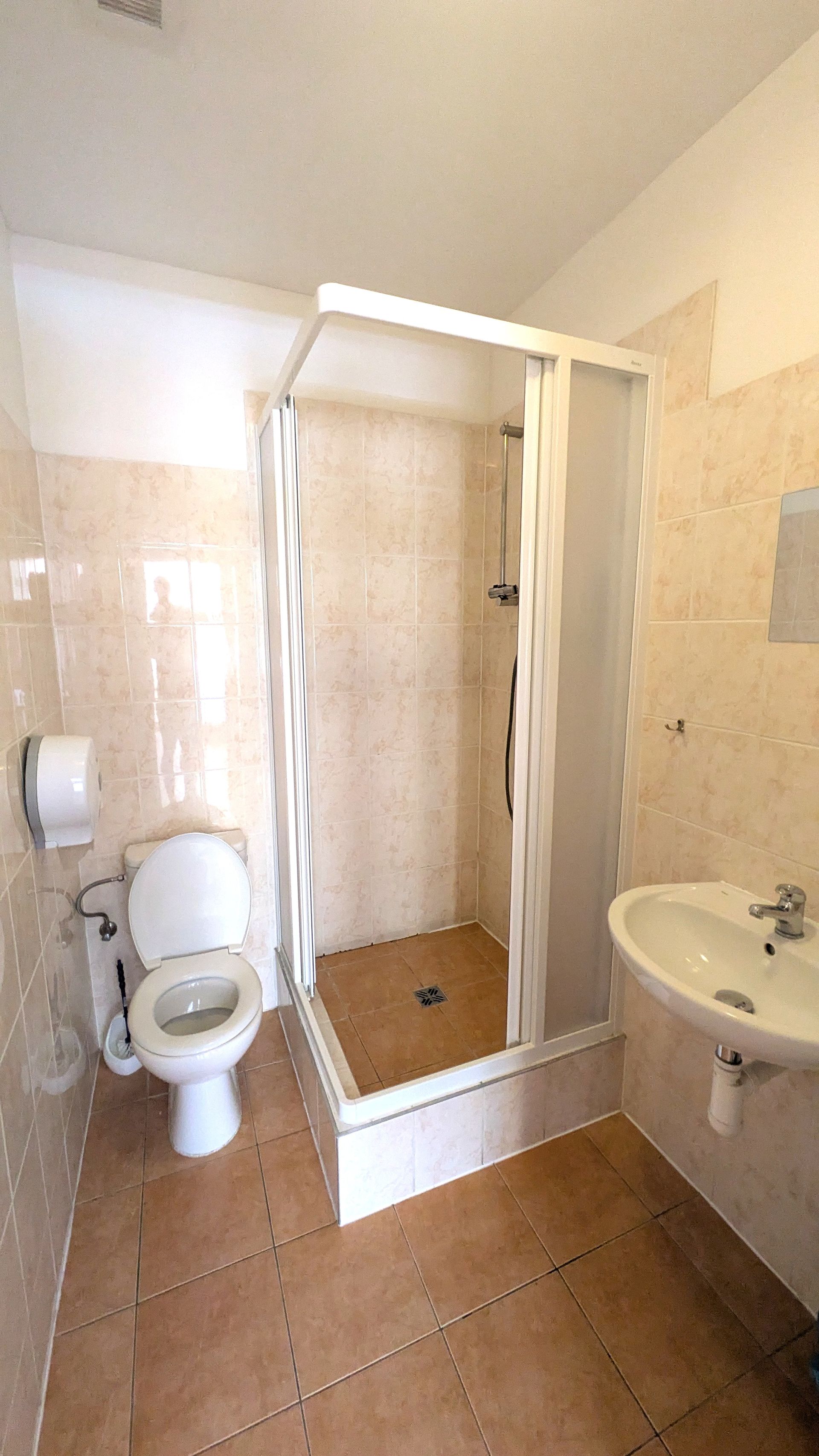 Bathroom with toilet, shower stall, and sink; tan tiled walls and floor.