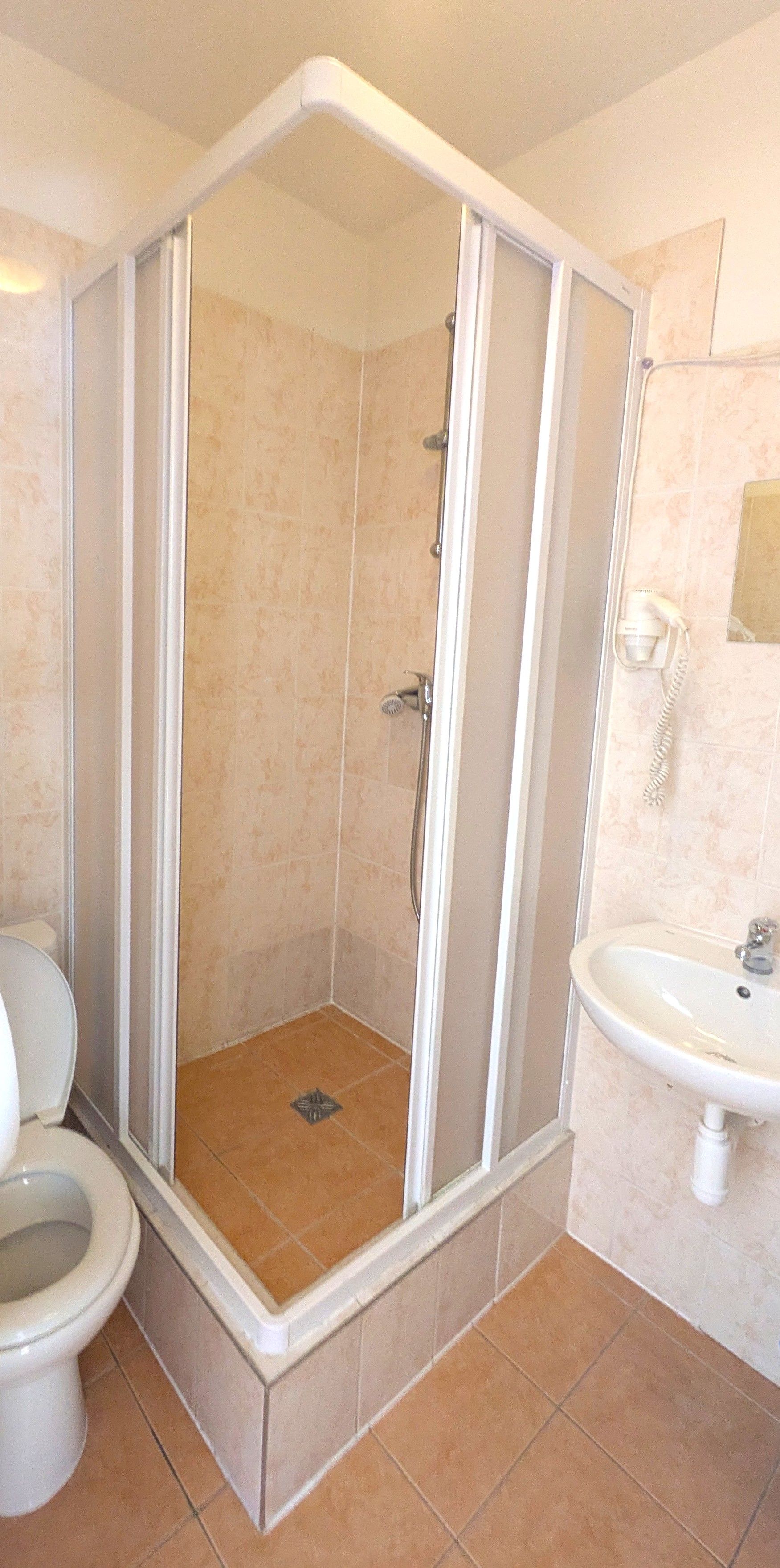 A bathroom featuring a corner shower, toilet, and a sink, with light brown tile throughout.