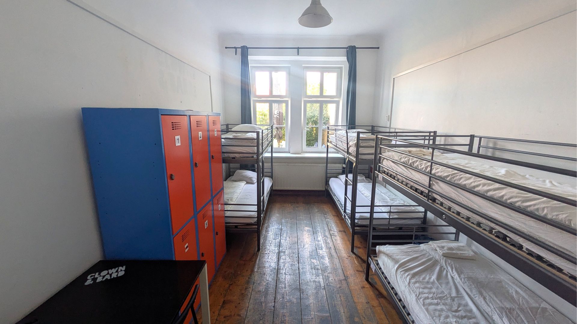 Hostel dorm room with three bunk beds, orange and blue lockers, window, and wooden floor.