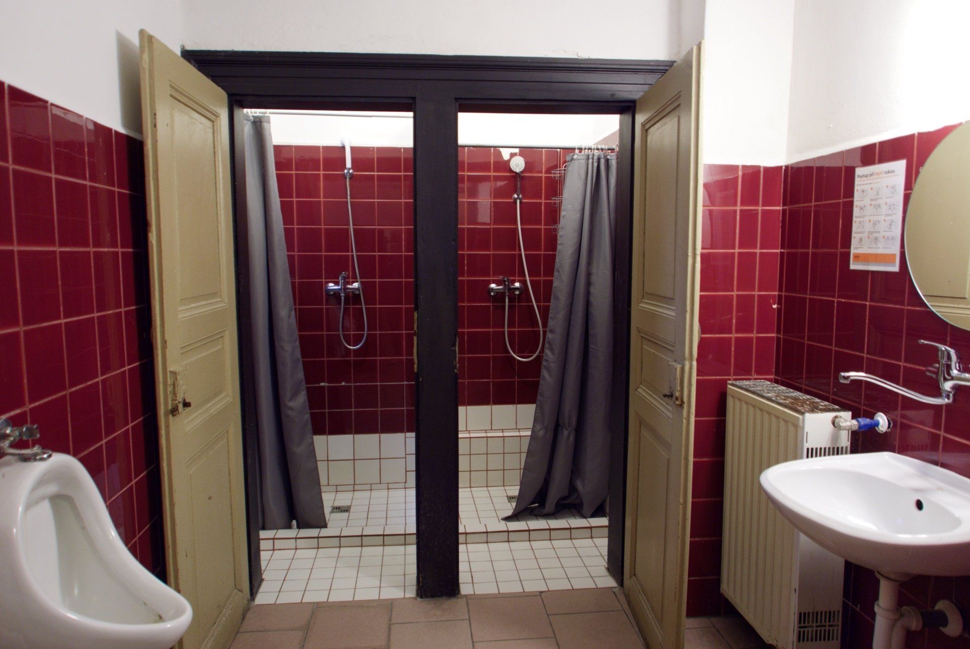 Public restroom with two shower stalls, red tile, gray curtains, and a urinal.