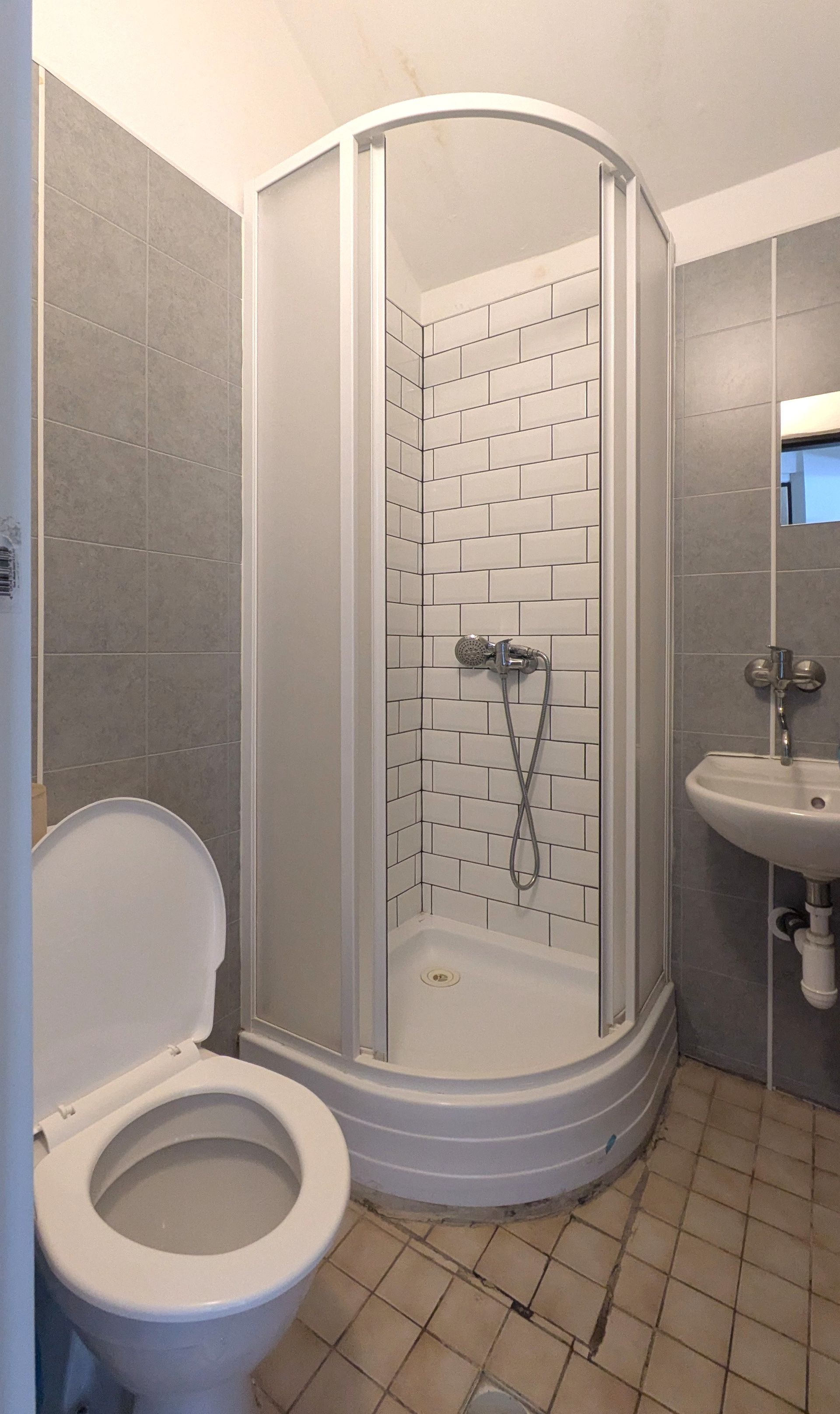 Small bathroom with toilet, curved shower stall, and sink against gray tiled walls.