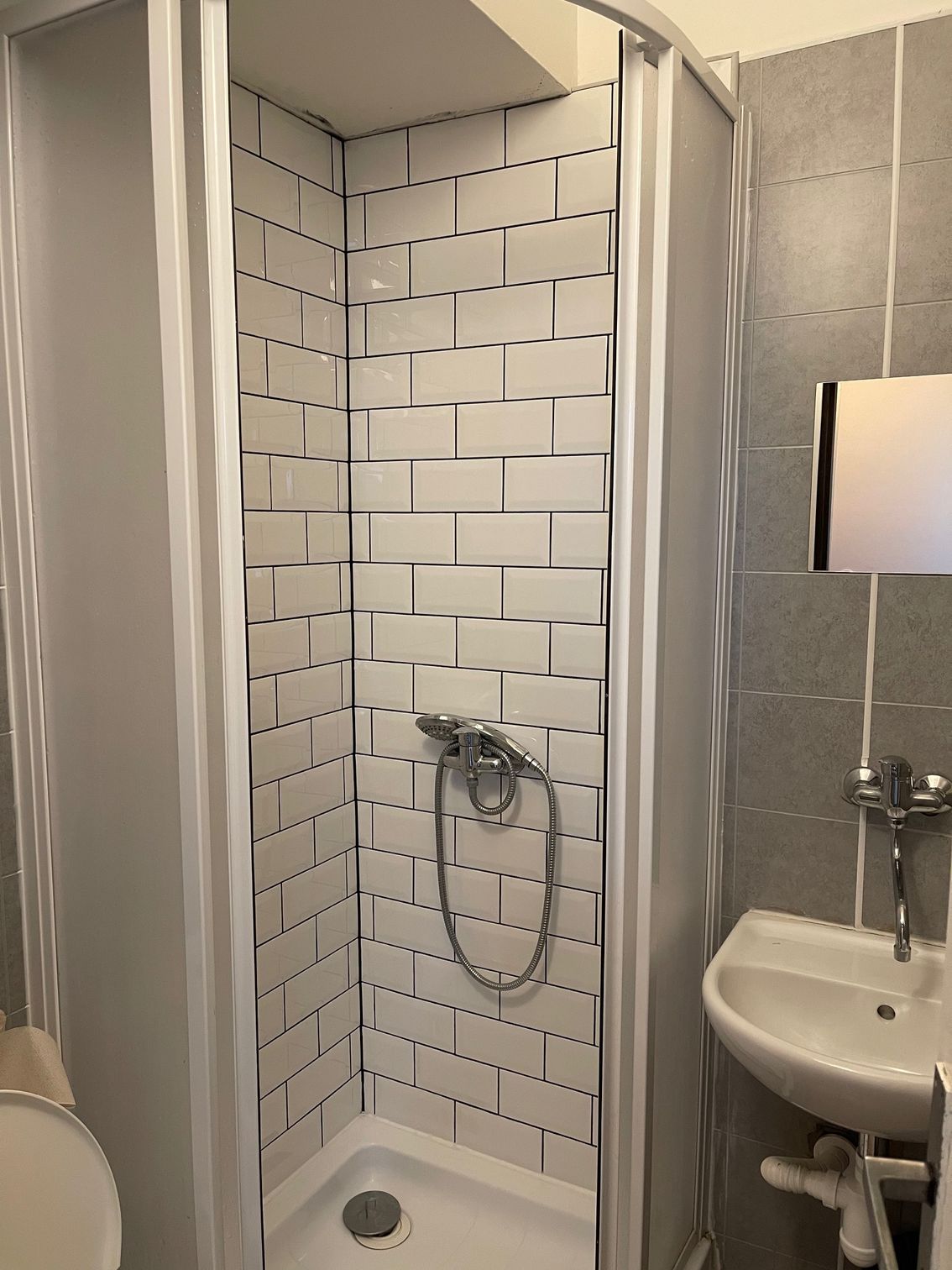 Shower stall with white brick-style tiles, silver shower head, and small sink in a bathroom.