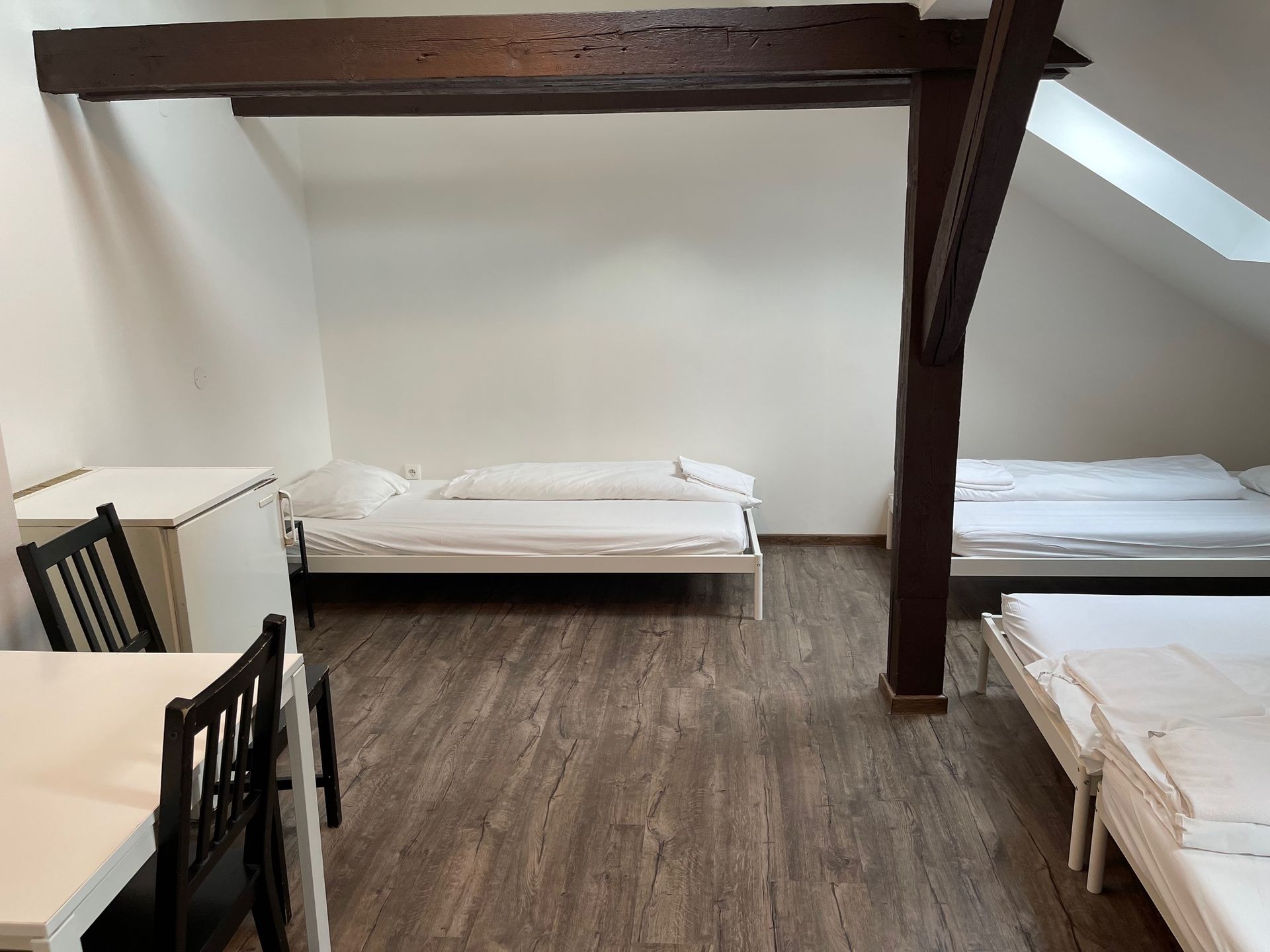 Attic bedroom with multiple beds, white walls, wood beams, and wooden floor. A table with chairs is on the left.
