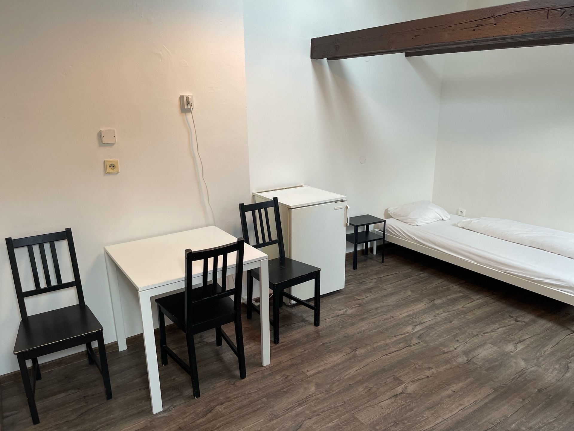 Room with bed, table, chairs, and small refrigerator against white walls and wood floor.