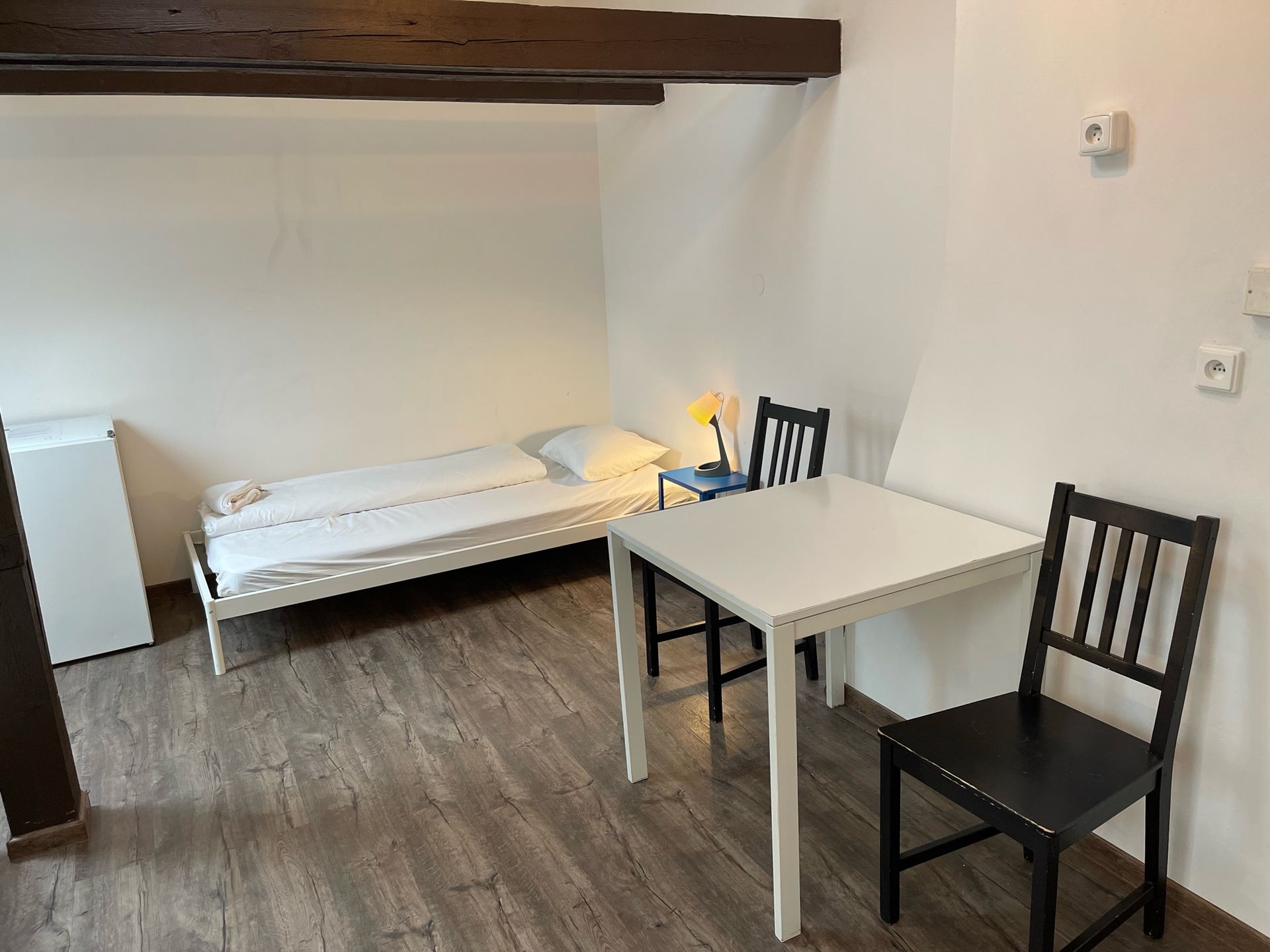 Room with a white bed, table, and two black chairs. Wooden floor and white walls.