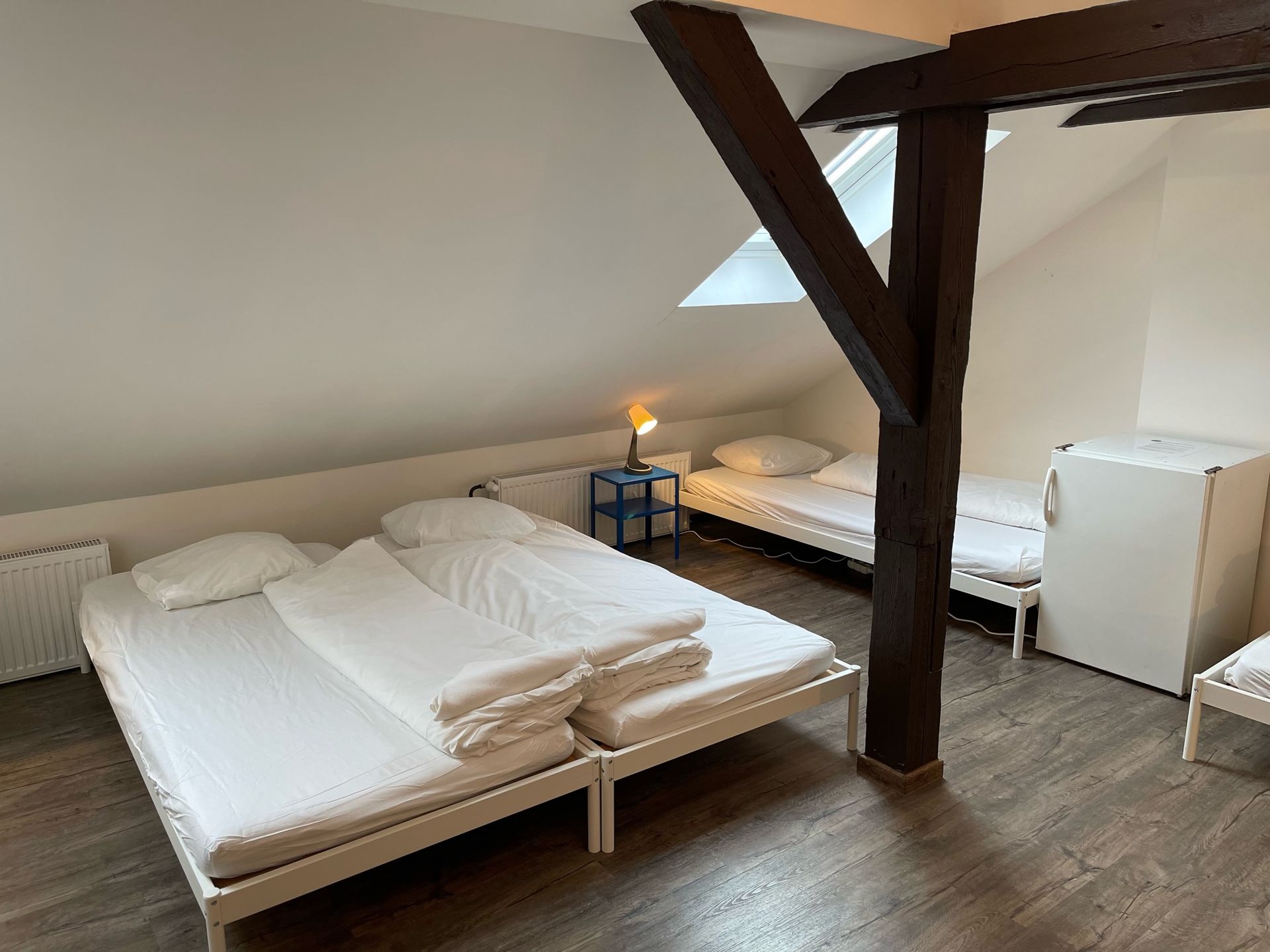 Attic bedroom with multiple beds, skylight, and dark wooden beams. White bedding and light wood floor.