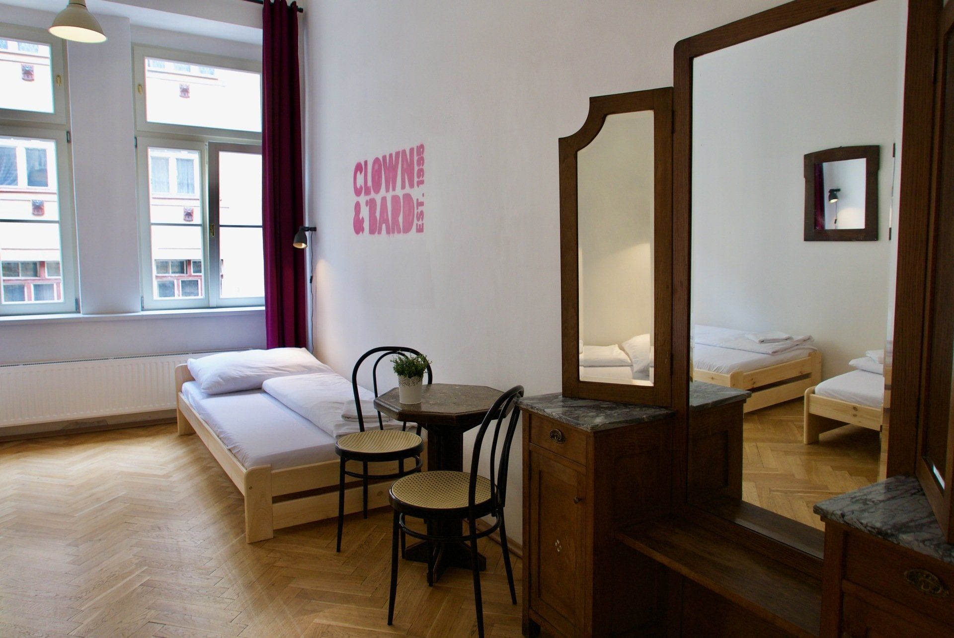 Bedroom with two beds, table, chairs, large mirror. White walls, wooden floor, windows.