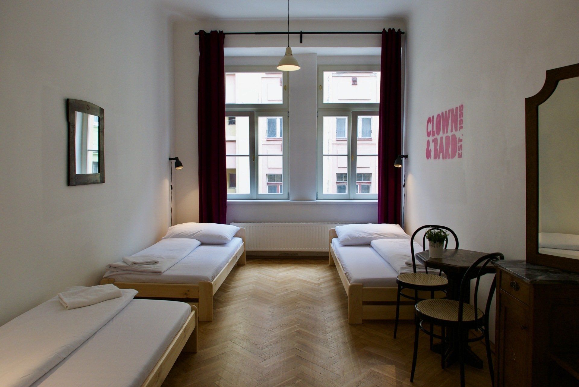 A hostel room with three beds, a small table, a mirror, and a window with red curtains.