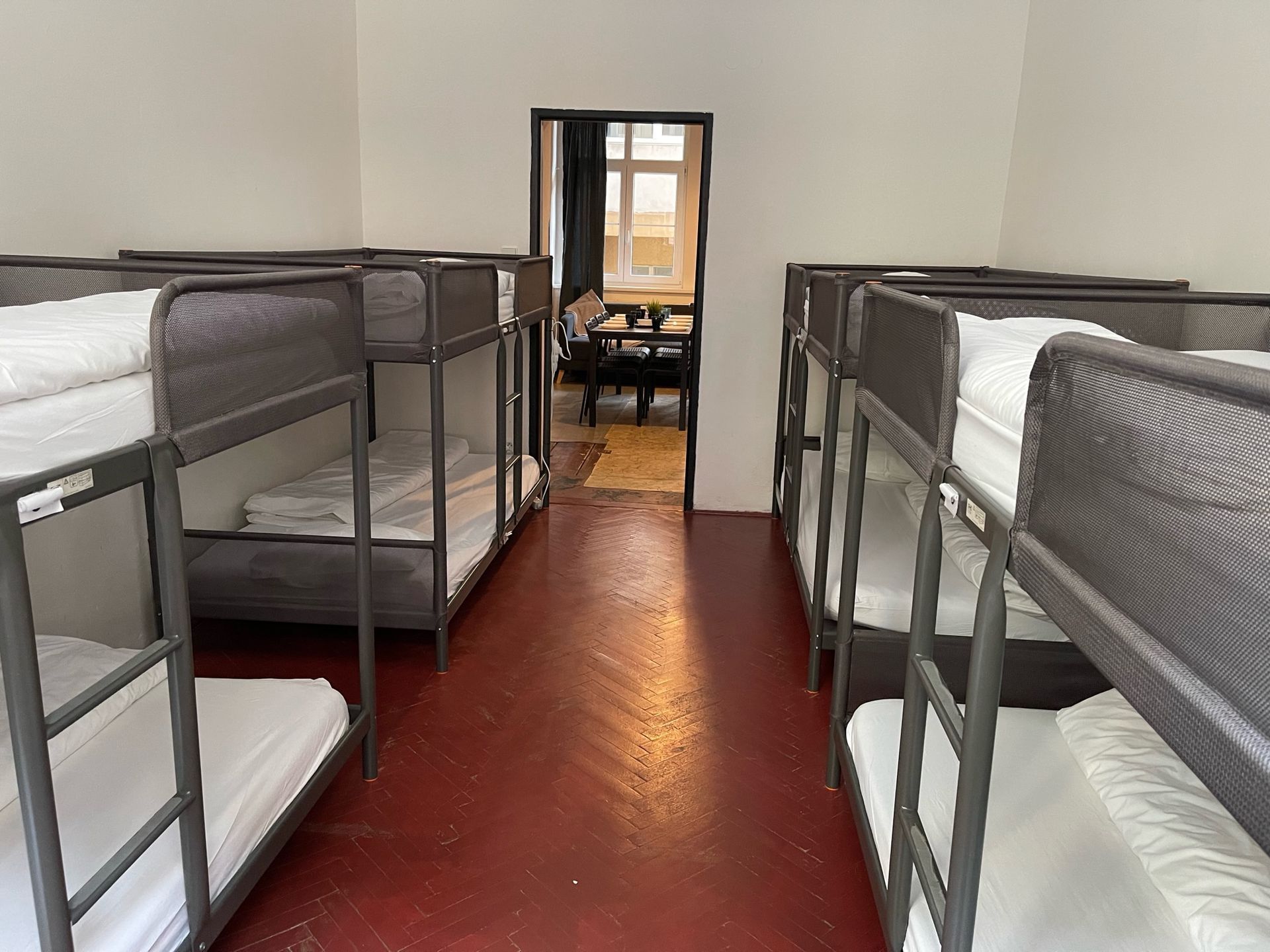 Dorm room with multiple bunk beds. Red floor. View through a doorway to a dining area with a table and chairs.