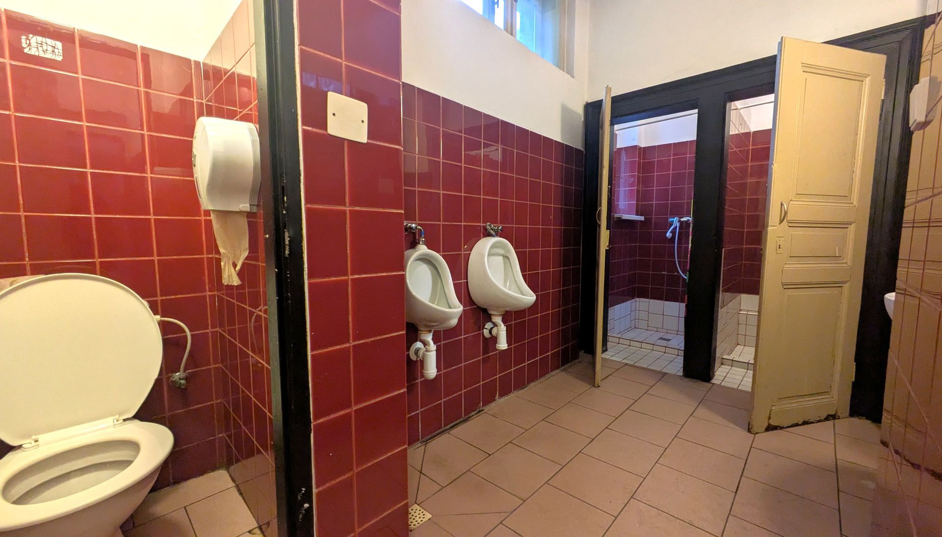 Public restroom with red tiled walls, two urinals, a toilet, and shower stalls.