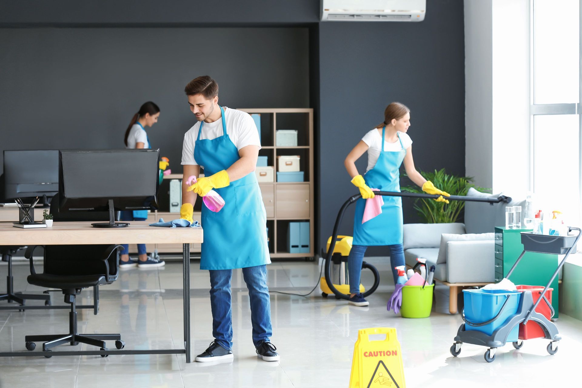 Office cleaners in blue aprons cleaning desks, vacuuming, and wiping windows.