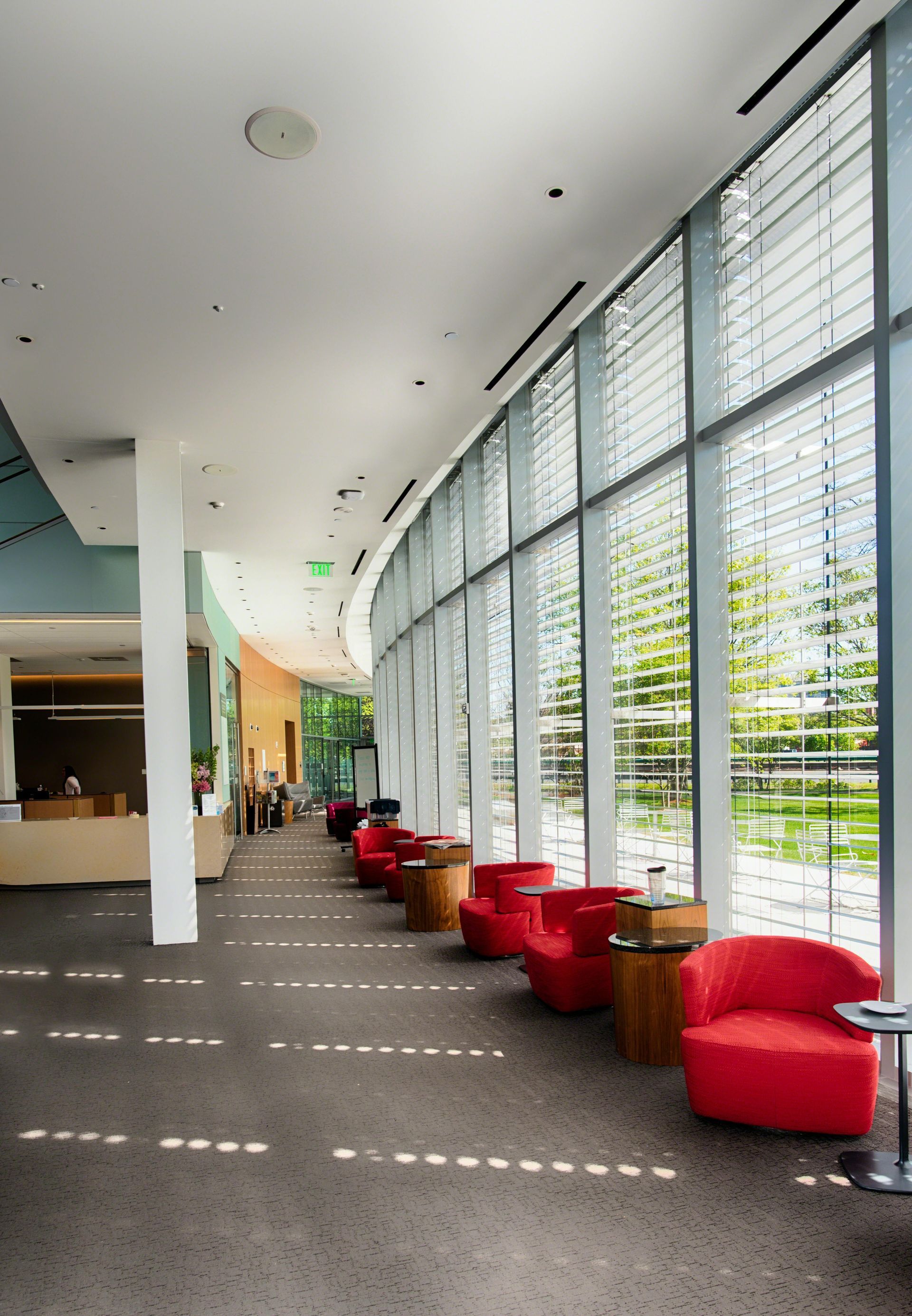 Curved hallway with red chairs, floor-to-ceiling windows with blinds, and white columns.