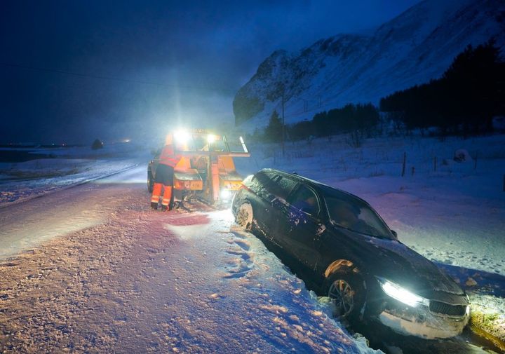 A car stuck in deep snow being pulled out by a small plow truck on a snowy road at night.