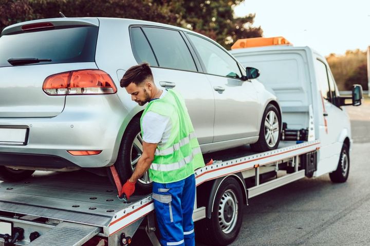 Tow truck operator securing a silver car on a flatbed tow truck. Outdoors, bright day.