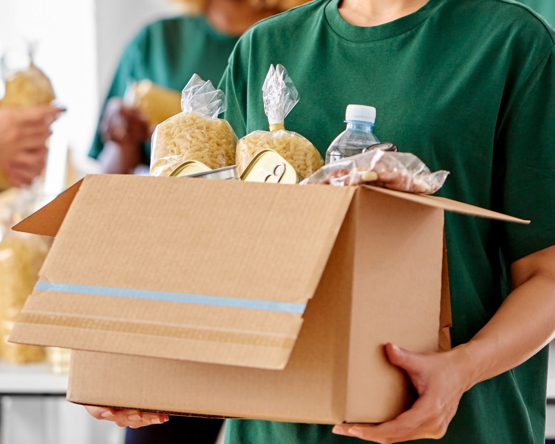 Person in green shirt carries a cardboard box filled with food for donation.