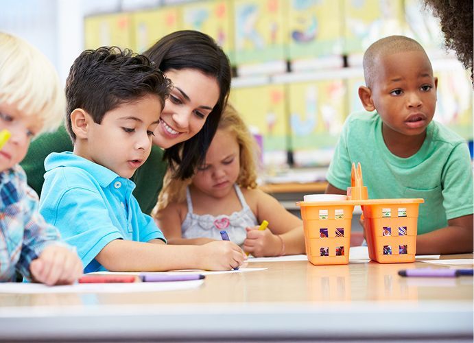 Teacher assists children drawing at a table in a classroom.