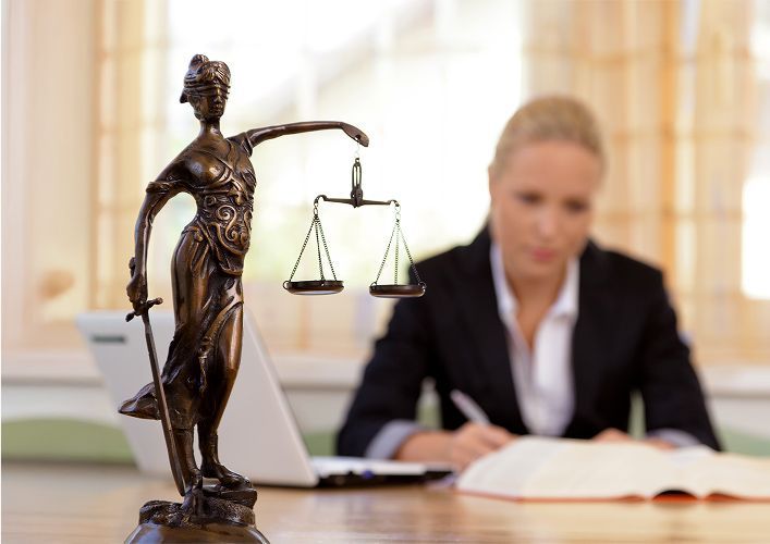 Statue of Lady Justice with scales, in front of a blurred businesswoman in a suit, working at a desk.