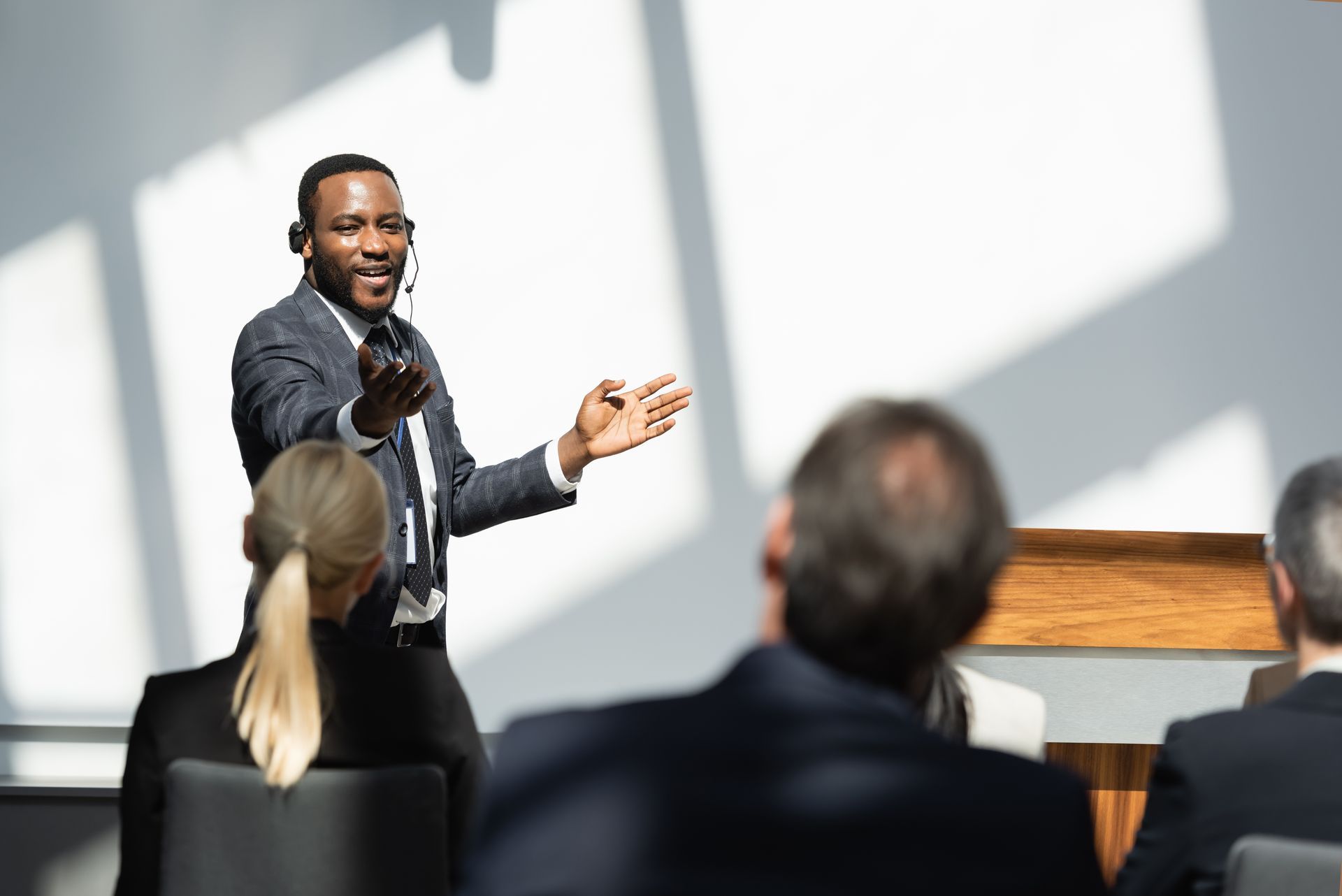 Man in suit gives presentation to audience; he gestures enthusiastically, wearing a headset microphone.