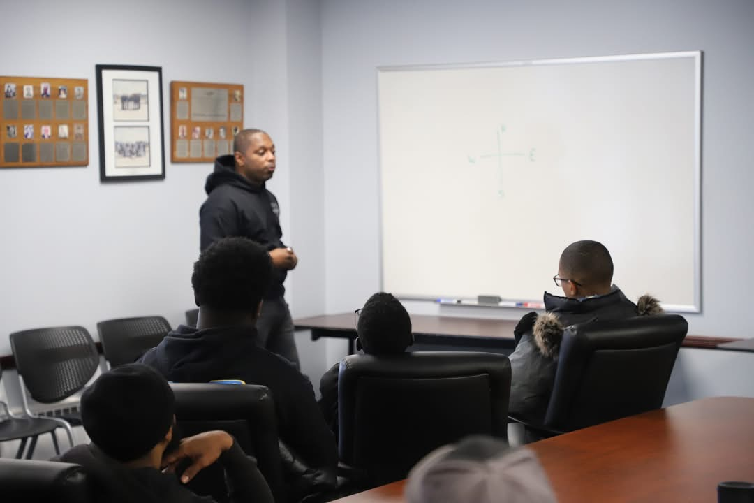 A man in black hoodie presents at a whiteboard to a small group of students in a classroom setting.