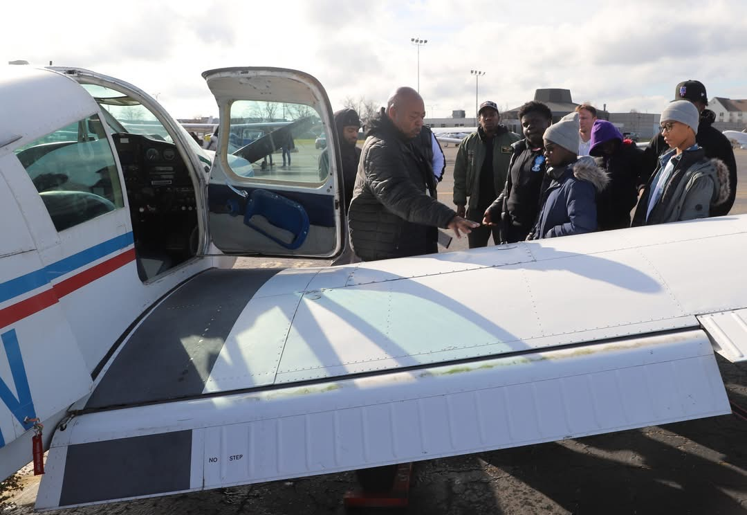 Group of people by a small airplane, one person shaking hands with another. Outdoors, sunny day.