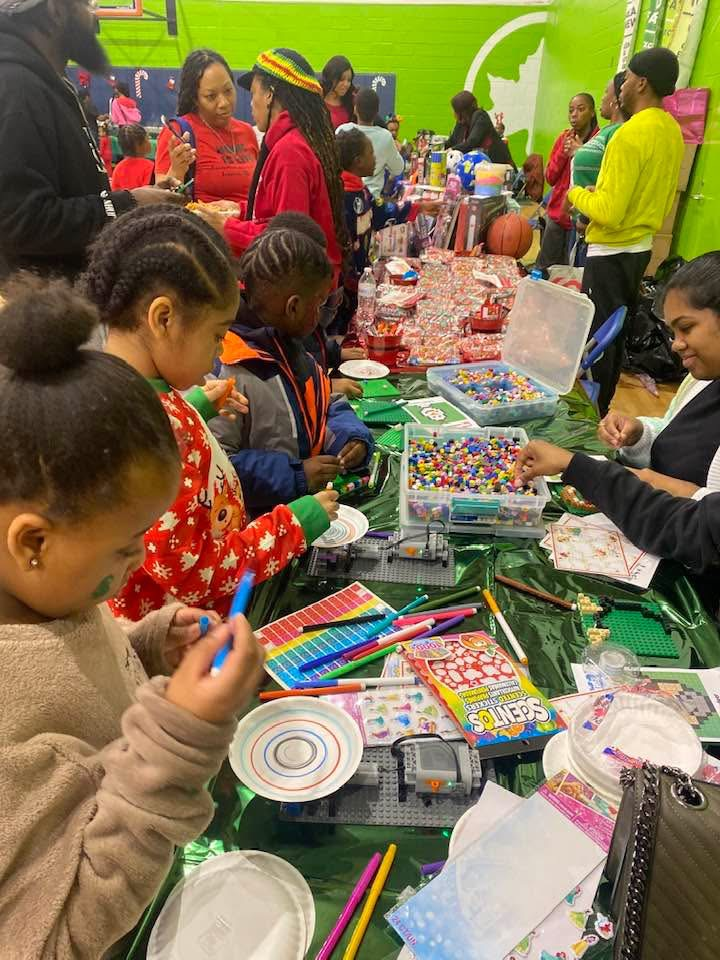 Children and adults craft at a table with beads and supplies. Indoors, green and red decor.