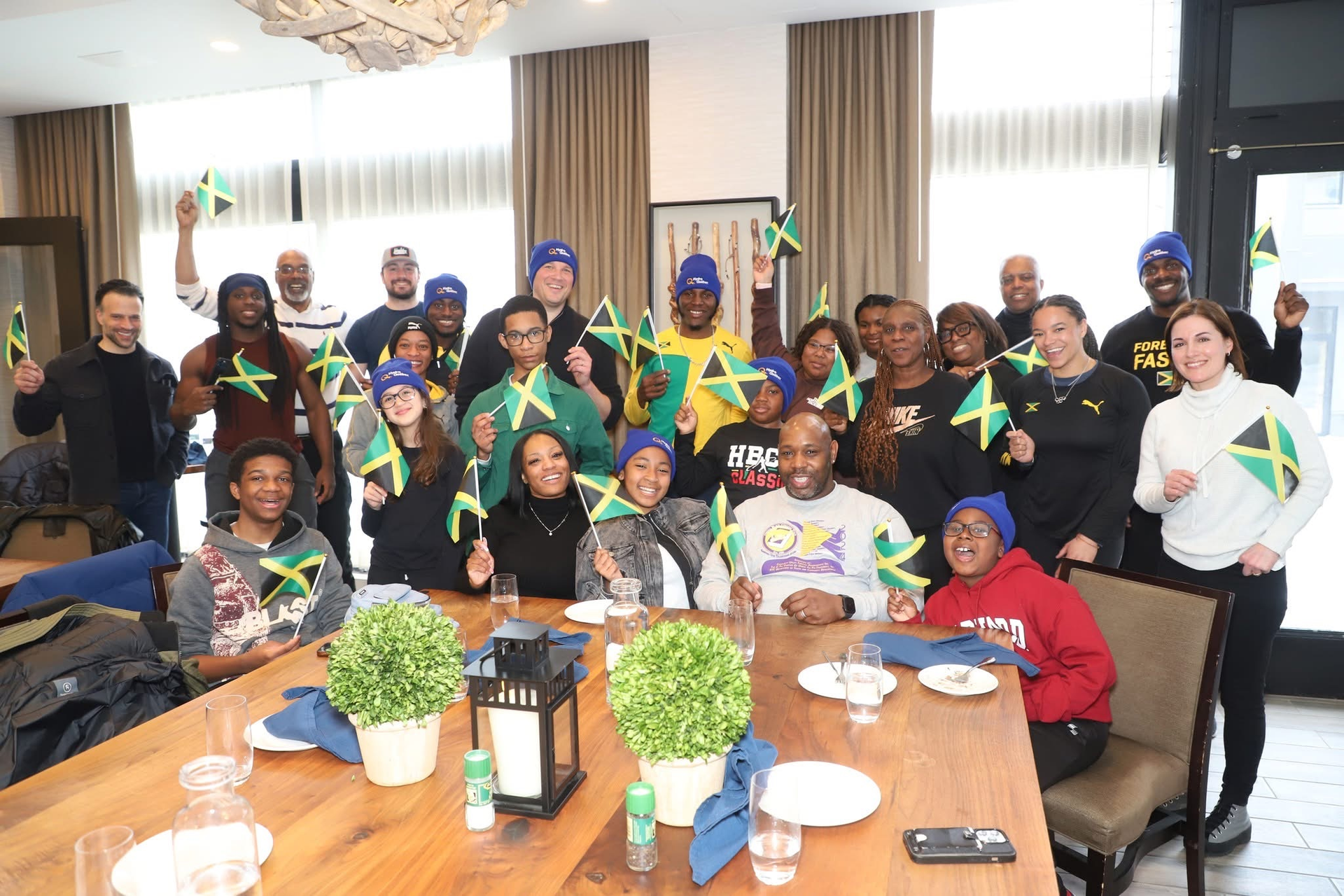 Group of people indoors, many holding Jamaican flags. Some wear blue hats, smiling.