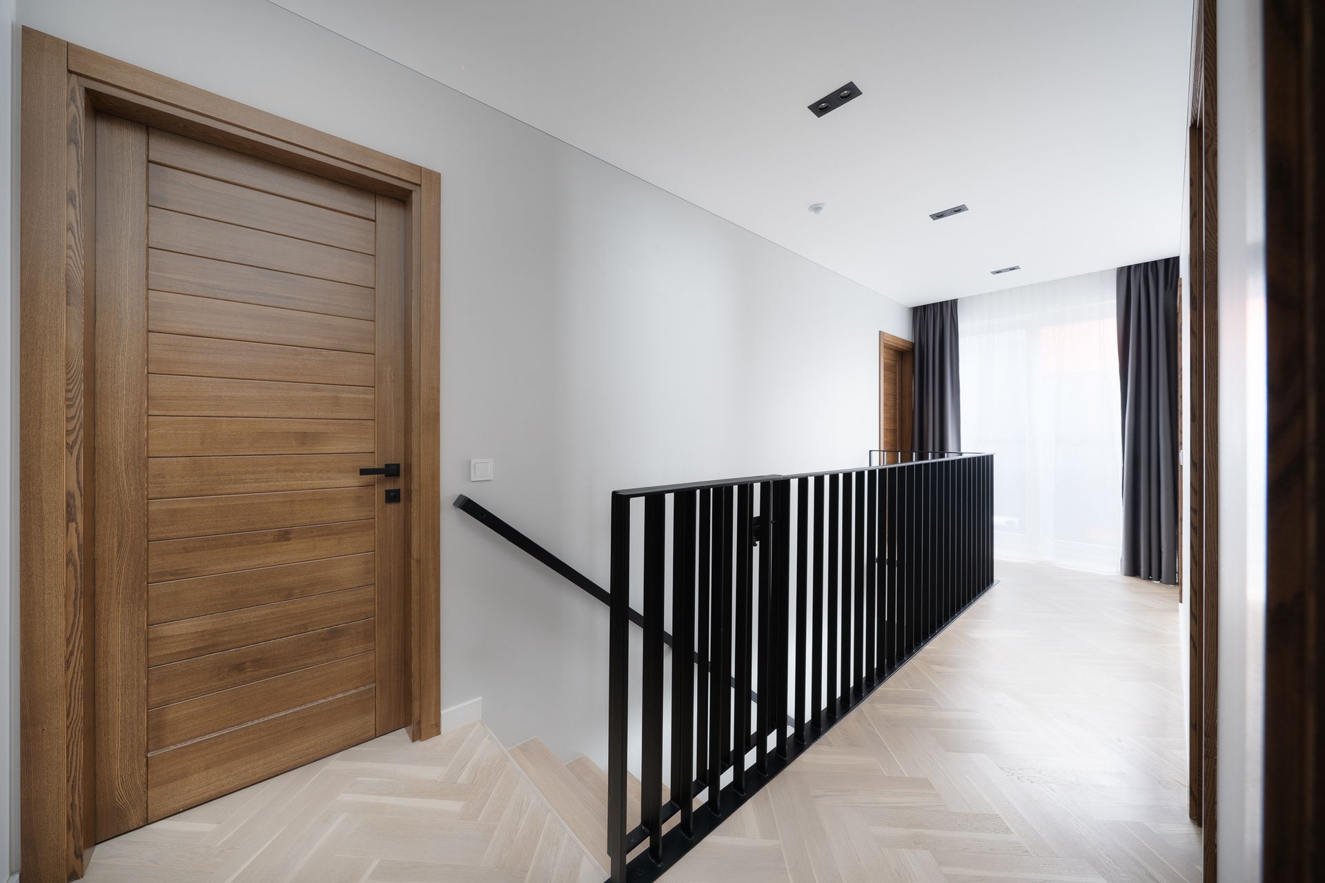 Koridorius su medinėmis durimis ir juodu turėklu, vedančiu į laiptus.