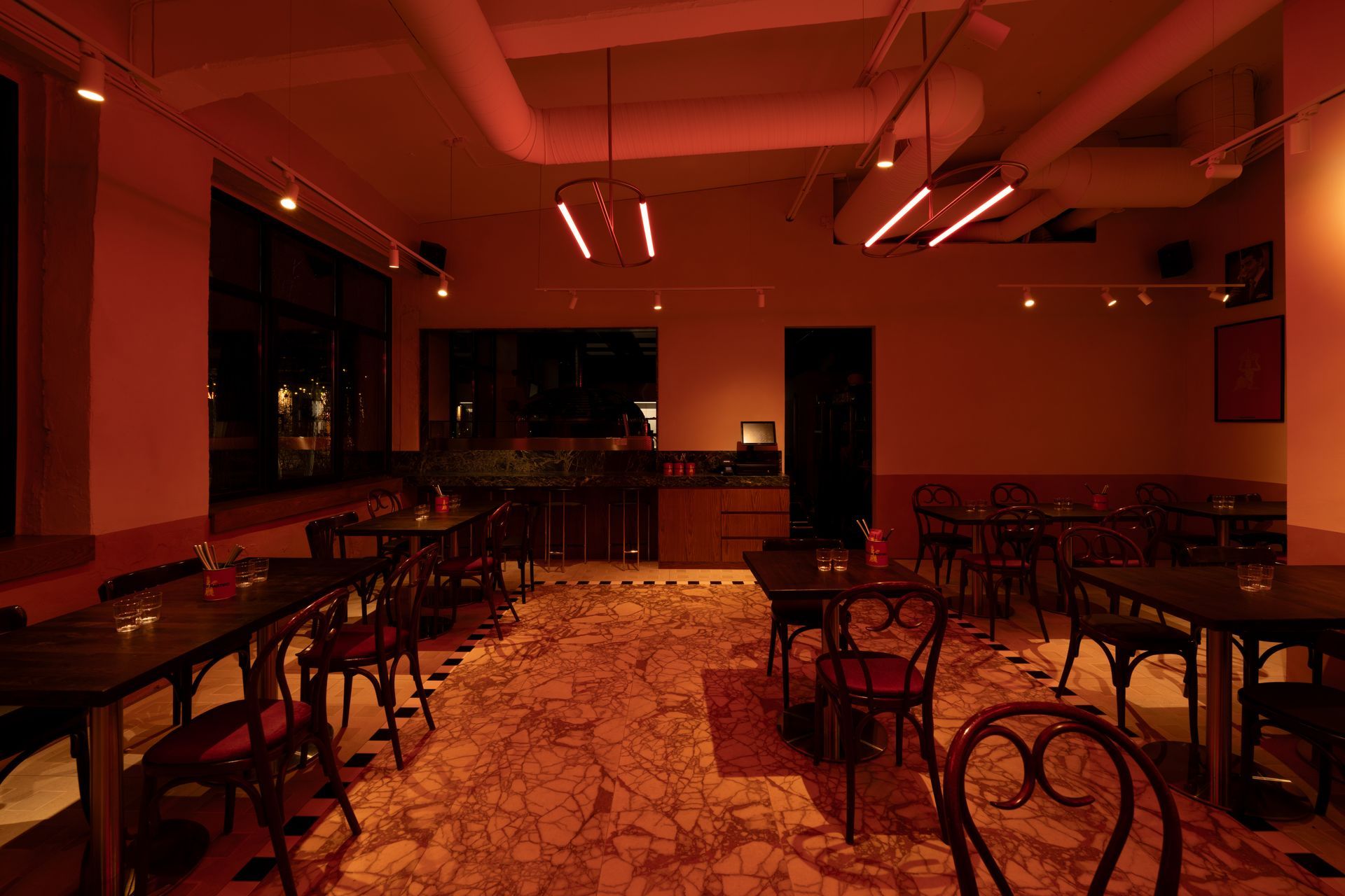 Empty restaurant interior with red lighting, tables, chairs, and patterned floor.