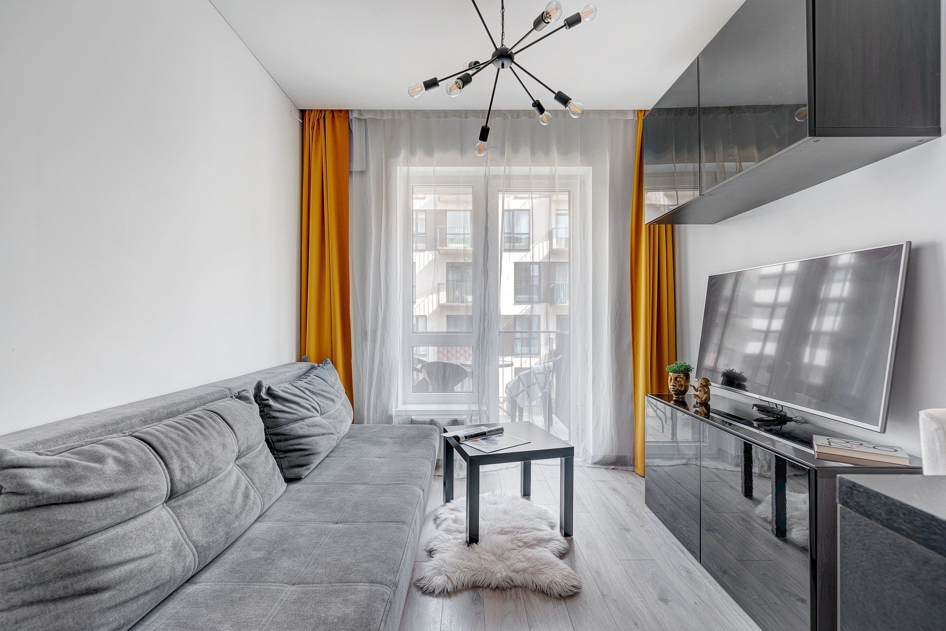 Svetainė su sofa, stalu, televizoriumi ir geltonomis užuolaidomis.