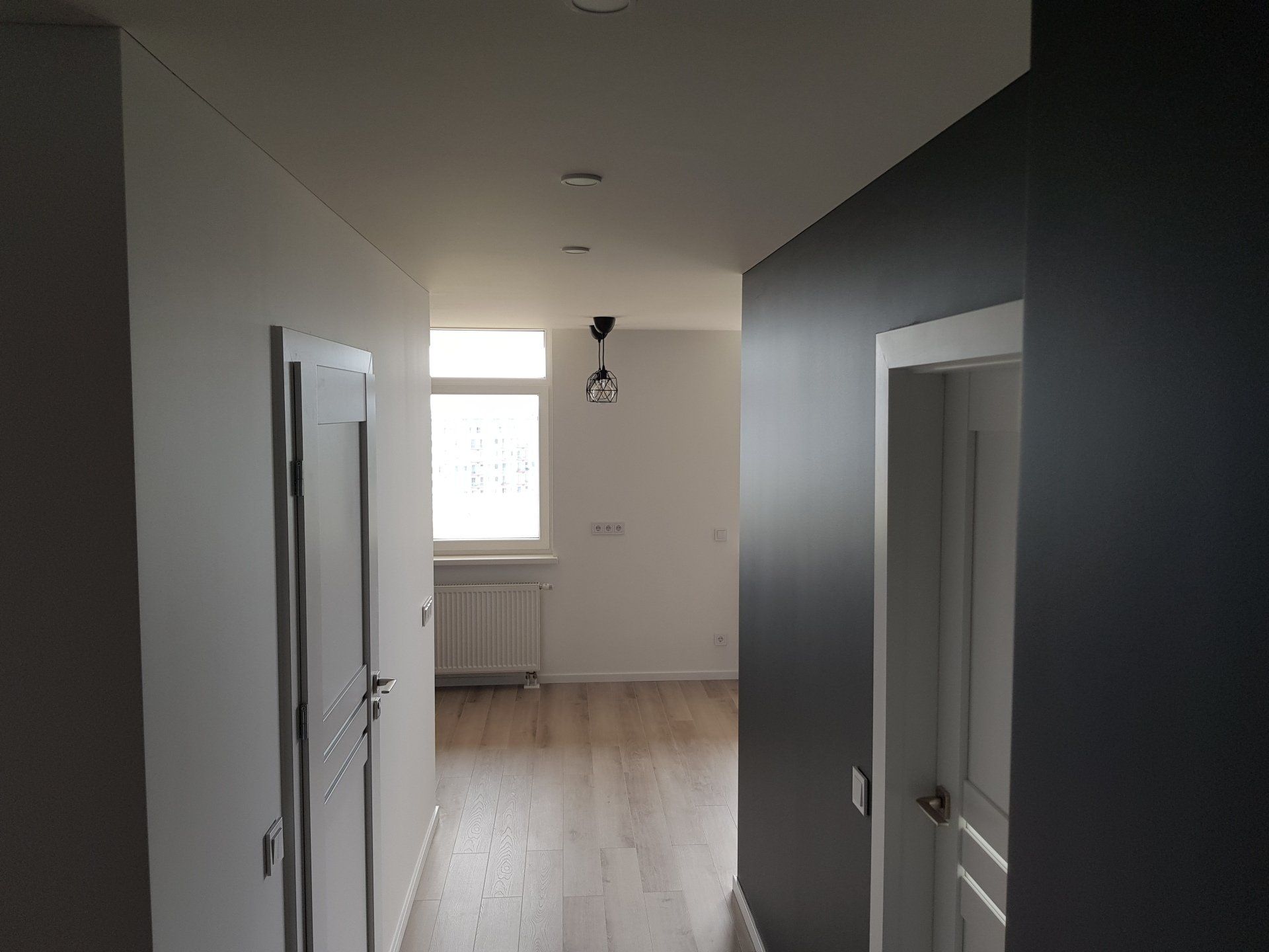 Koridorius su baltomis sienomis ir medinėmis grindimis, vedantis į svetainę.