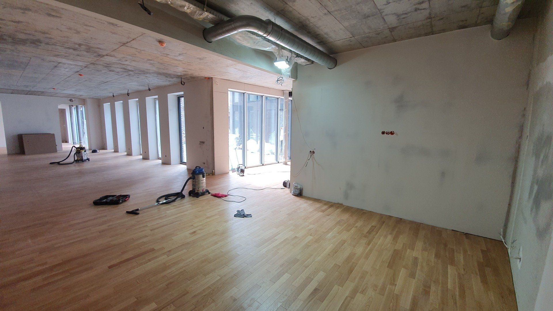 Tuščias kambarys su medinėmis grindimis ir ant grindų padėtu dulkių siurbliu.