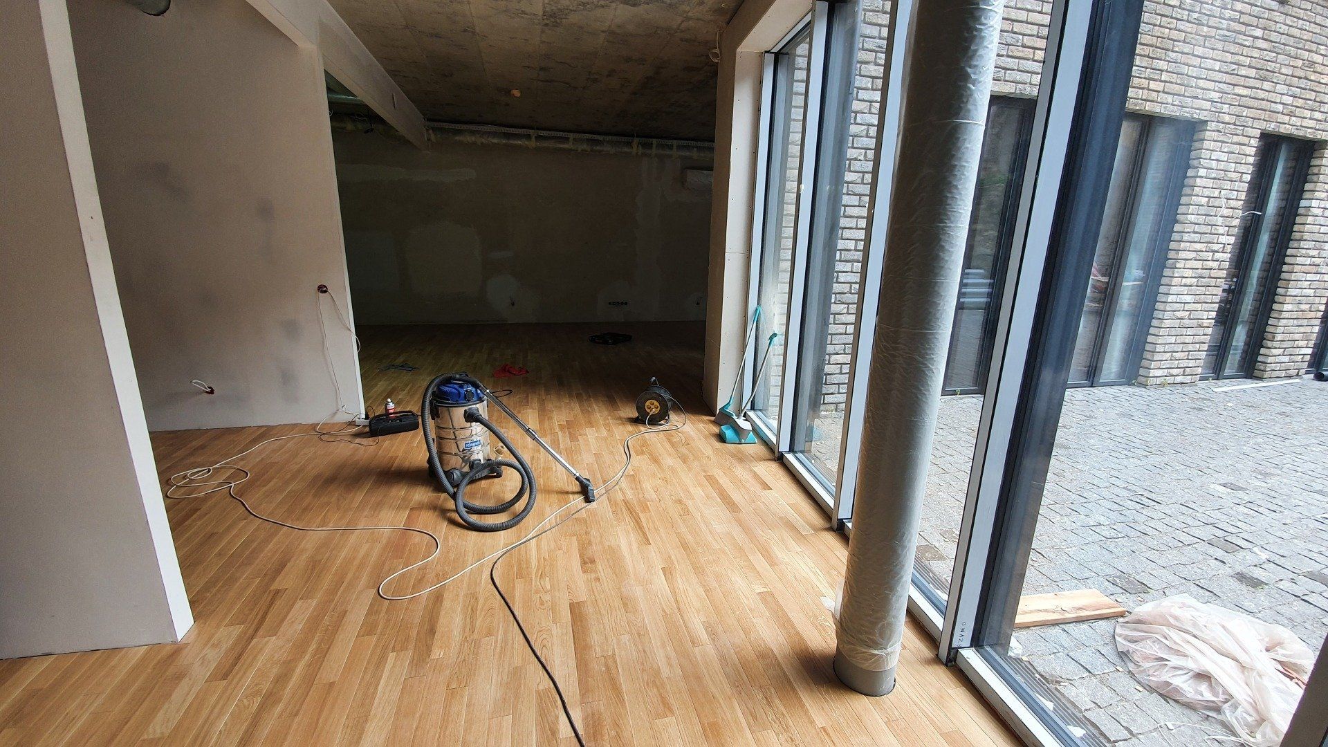 Kambaryje su daugybe langų ant medinių grindų stovi dulkių siurblys.