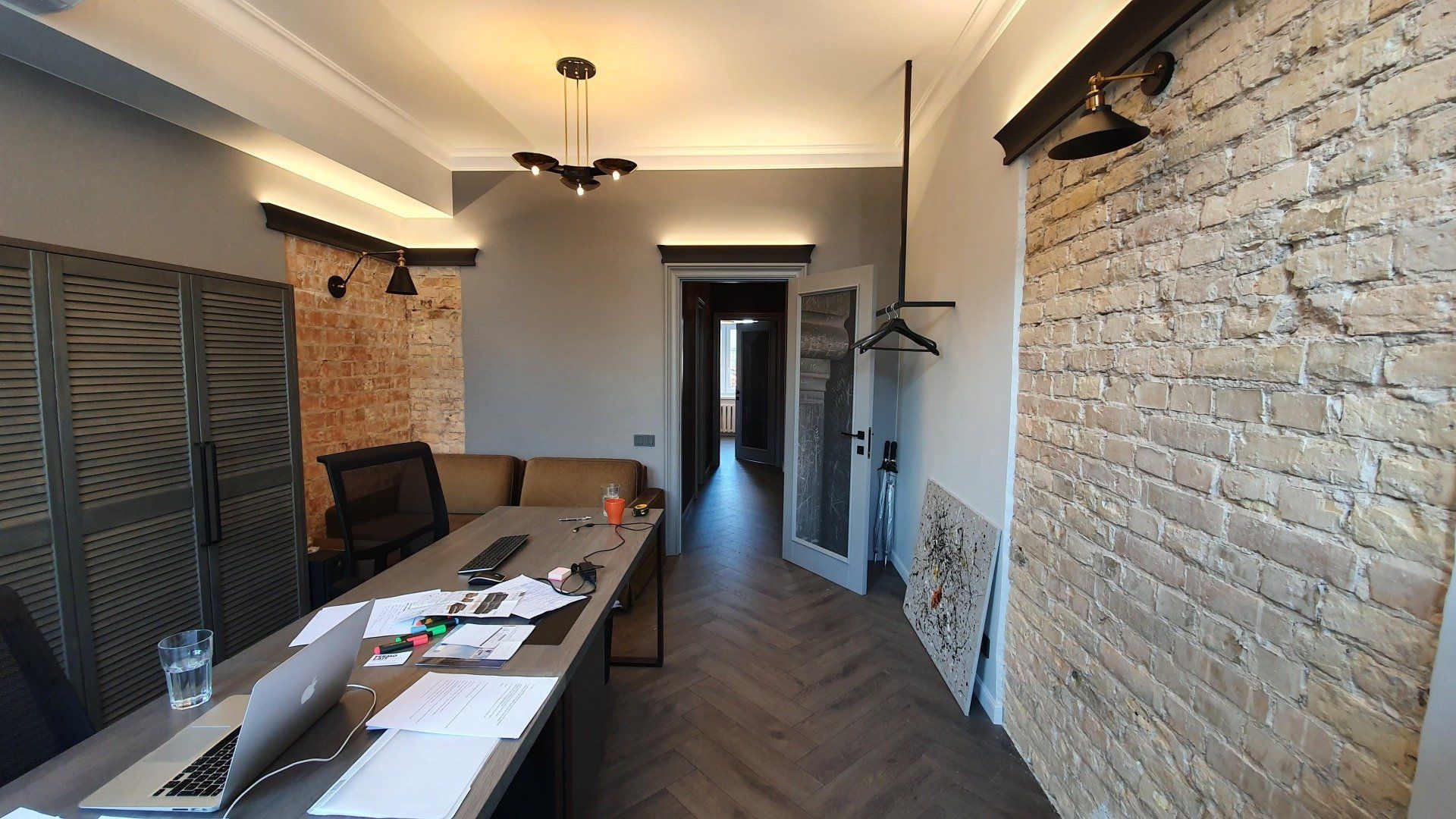 Ilgas koridorius su plytų siena ir stalu, ant kurio padėtas nešiojamas kompiuteris.