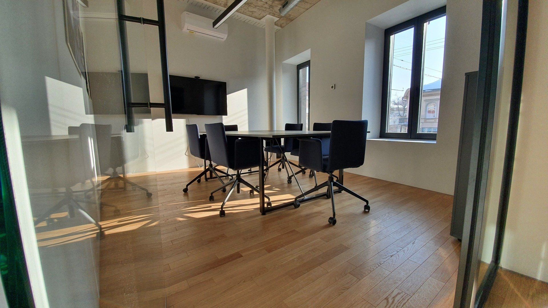 Konferencijų salė su stalu, kėdėmis ir televizoriumi.