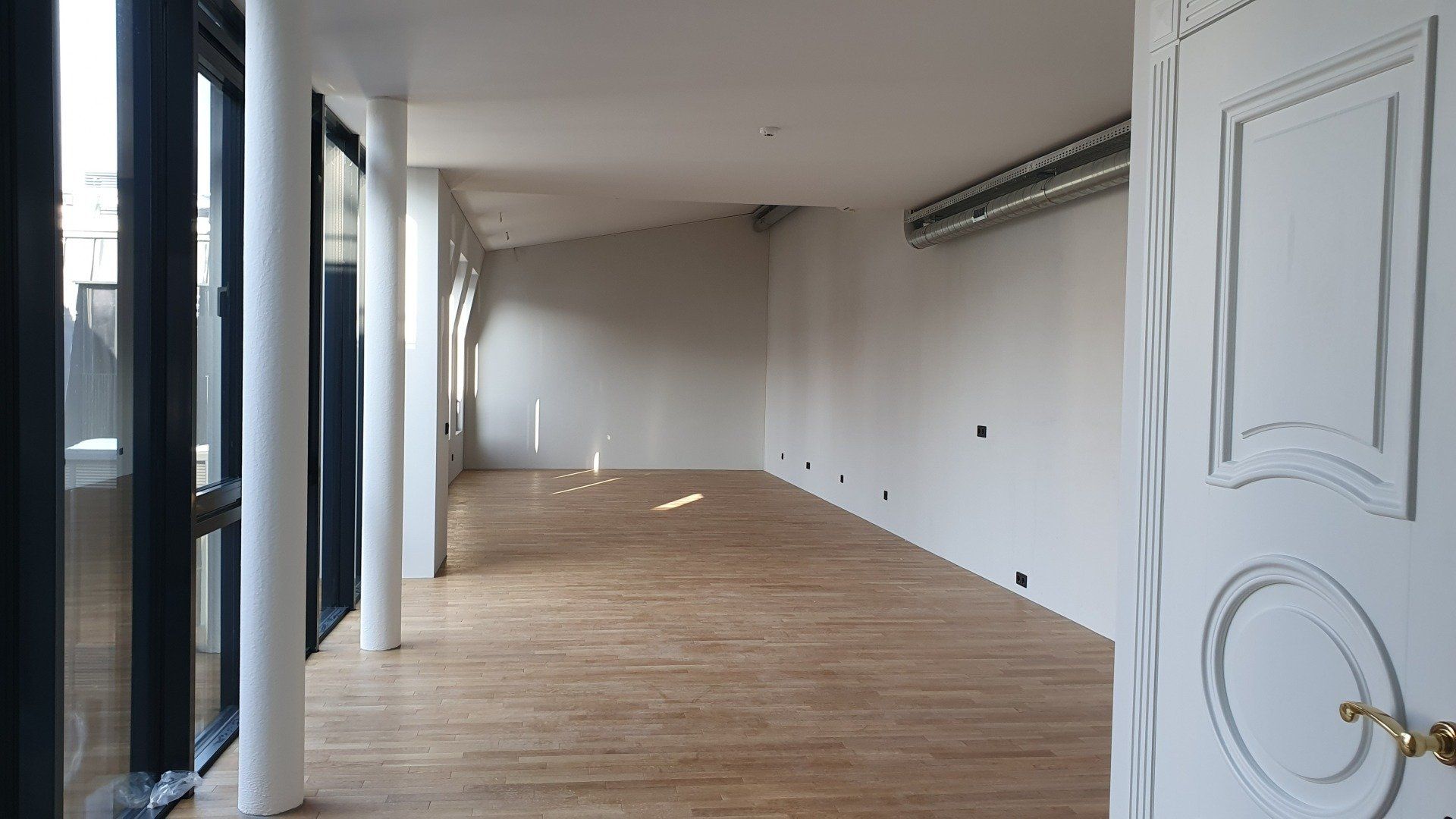 Didelis tuščias kambarys su medinėmis grindimis ir baltomis durimis.