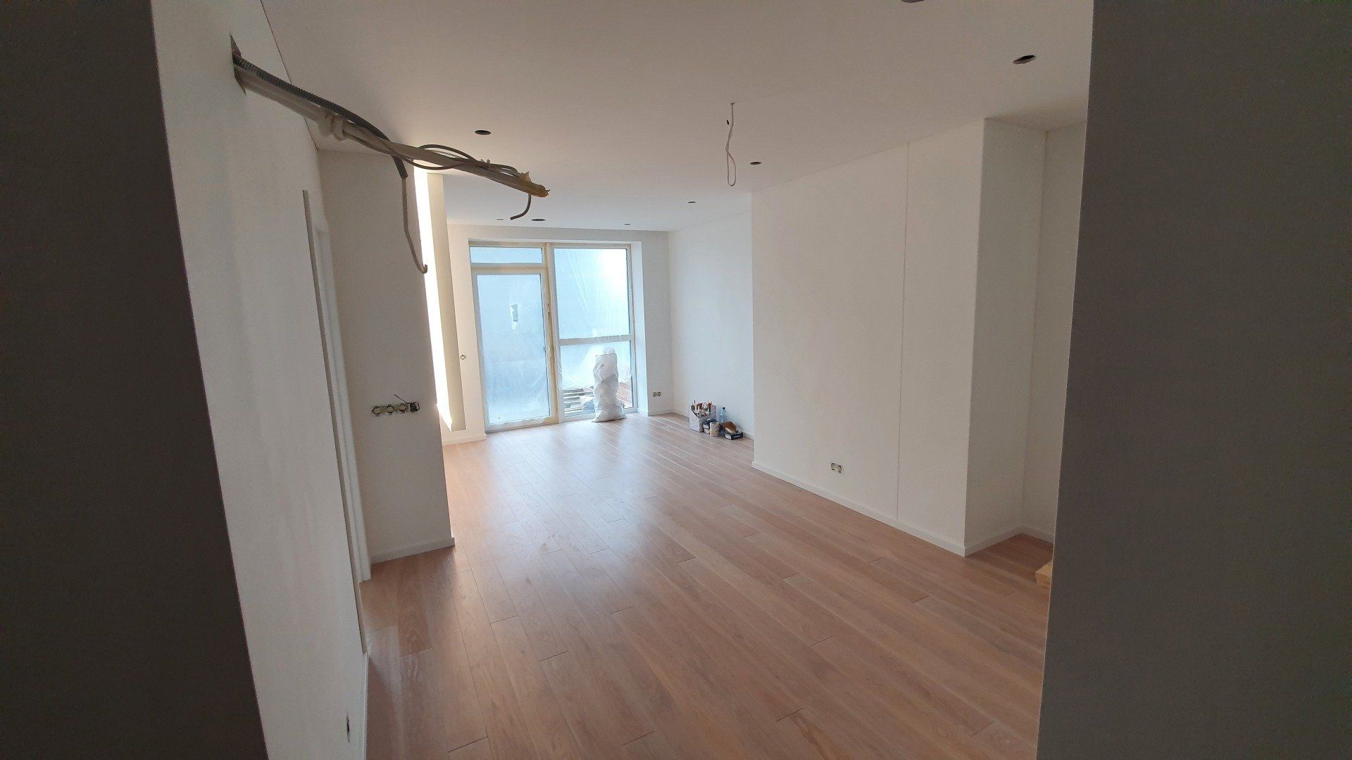 Didelis tuščias kambarys su medinėmis grindimis ir baltomis sienomis.