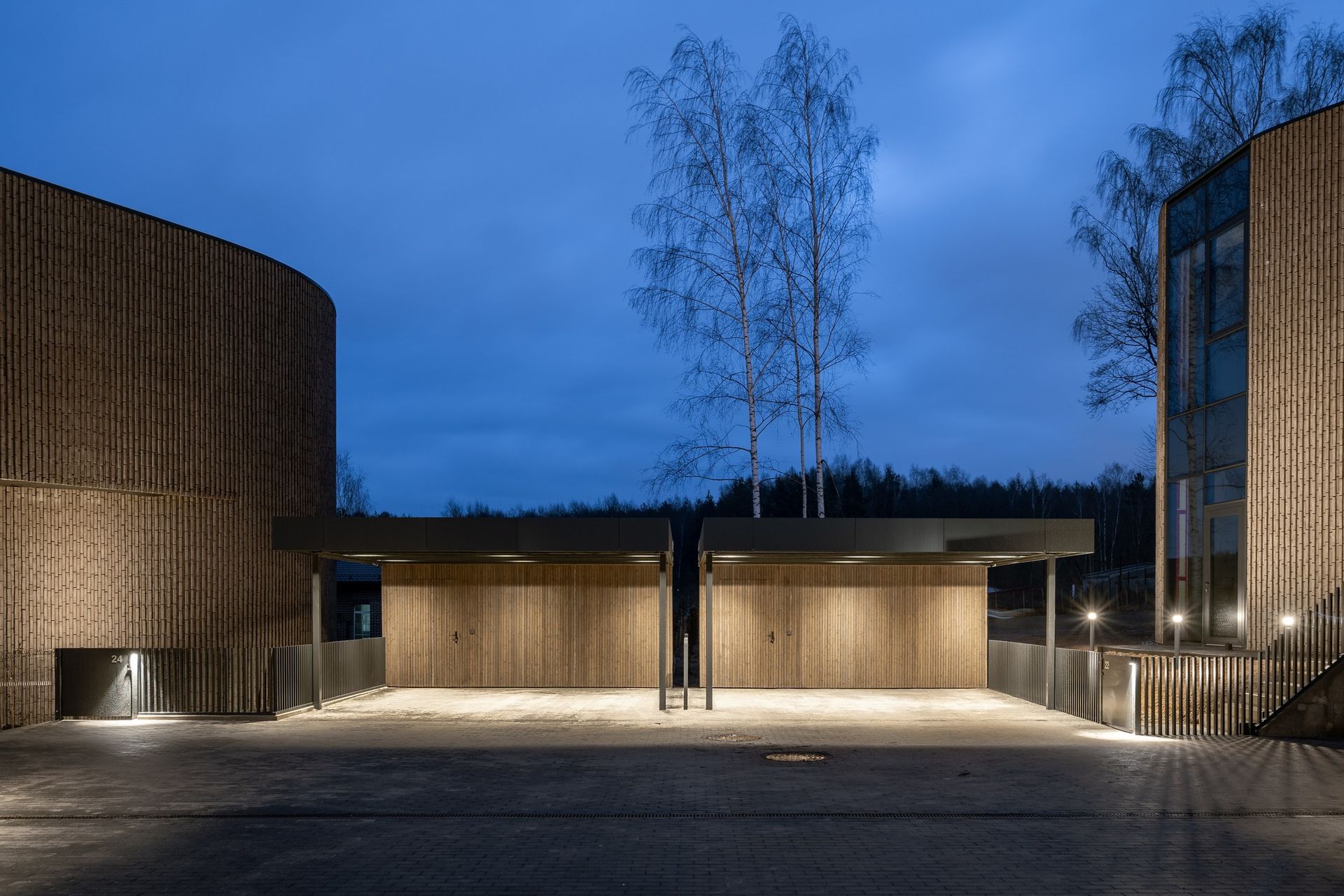 Modernus pastatas su mediniais garažo vartais ir medžiais priešais jį naktį.