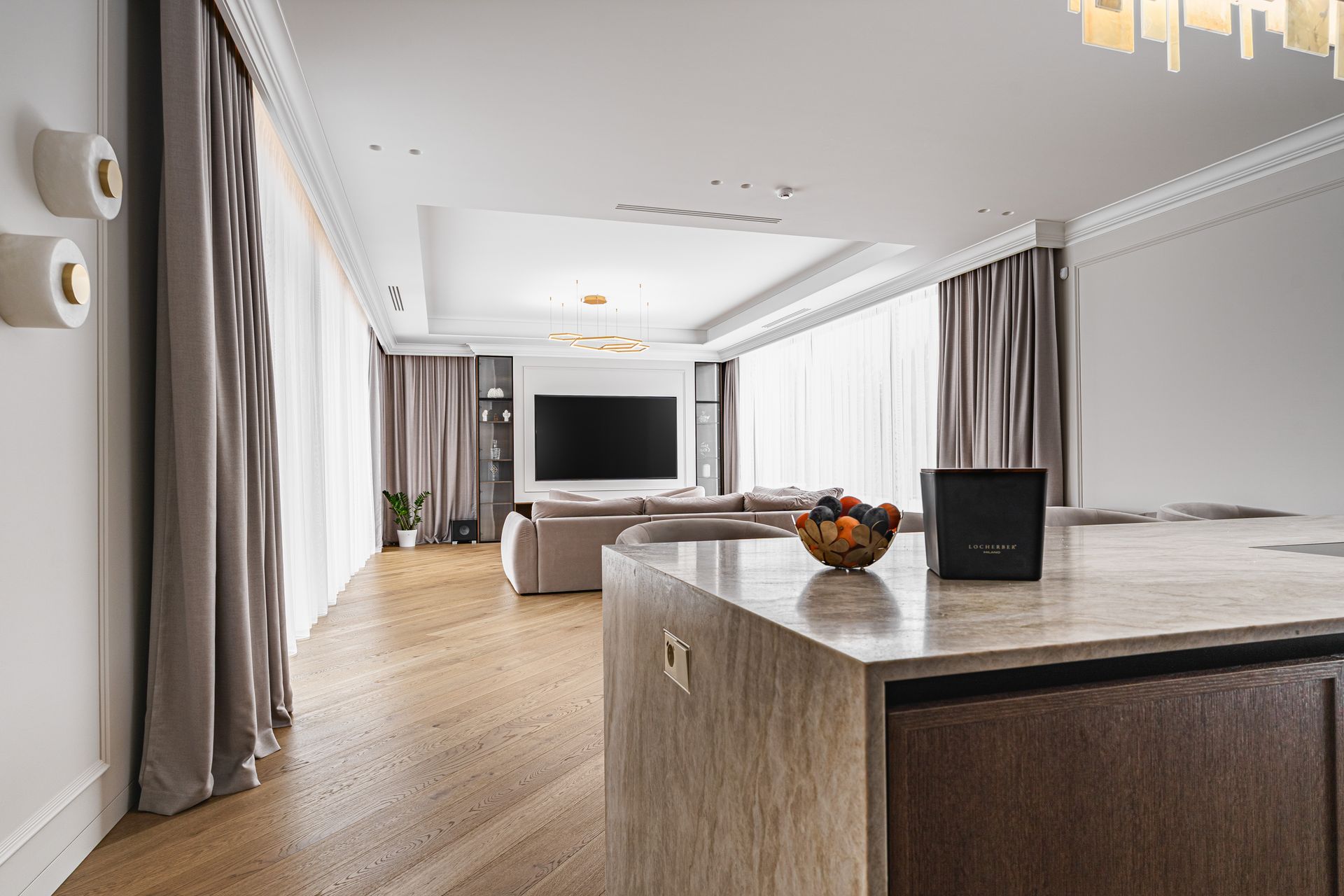 Spacious living room with large TV, curved island, and light wood floors. Gray curtains and white ceiling.