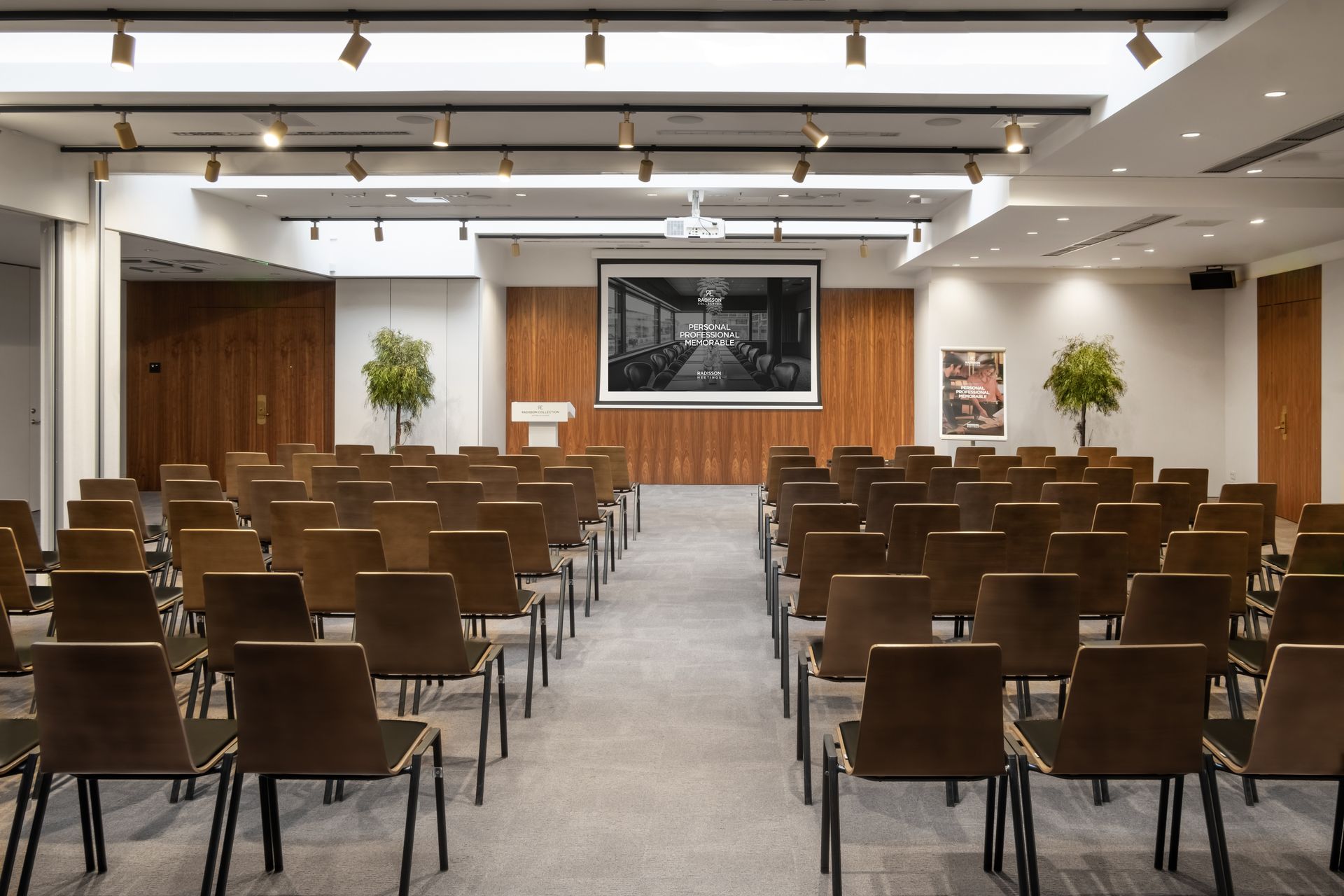 Didelė konferencijų salė su kėdžių eilėmis ir projektoriaus ekranu.