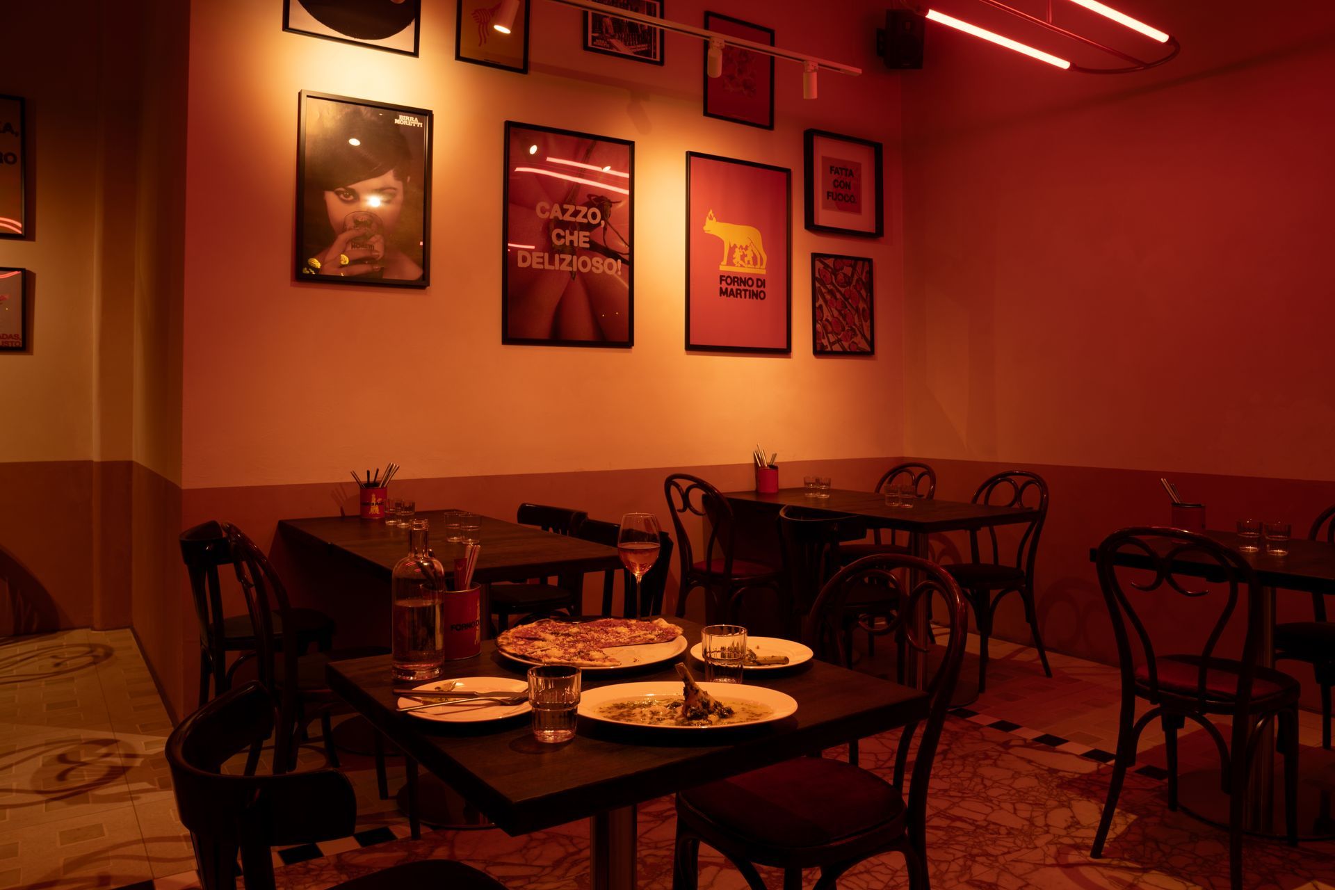 Interior of a restaurant with tables, chairs, and posters on the walls. Pizza and drinks on one table, illuminated by red light.