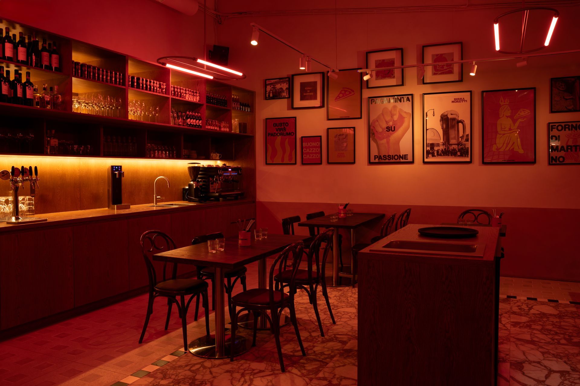 Red-lit bar interior with tables, chairs, shelving with bottles, and framed art.