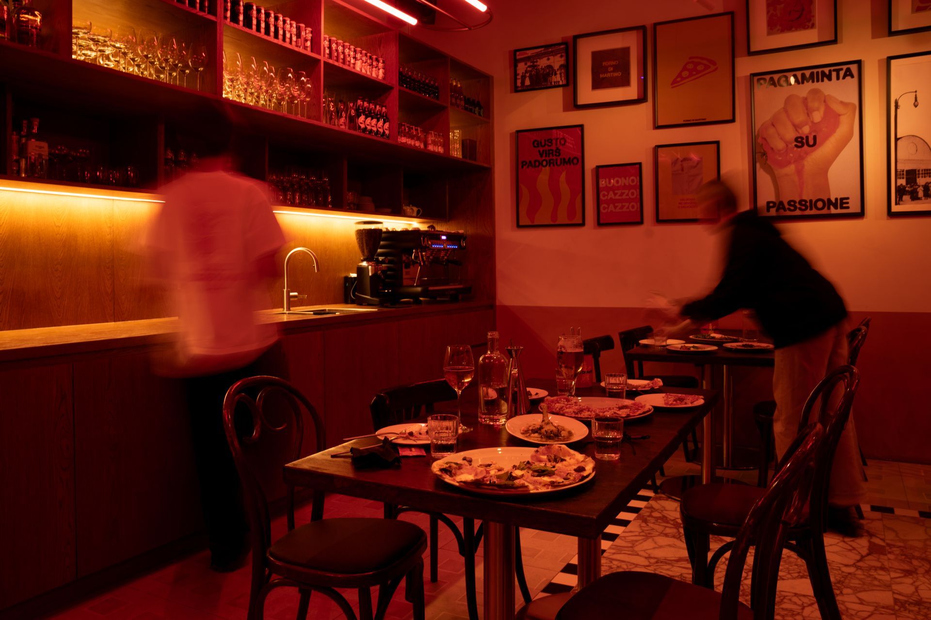Interior of a dimly lit restaurant with tables set. Servers move behind a bar illuminated in red.