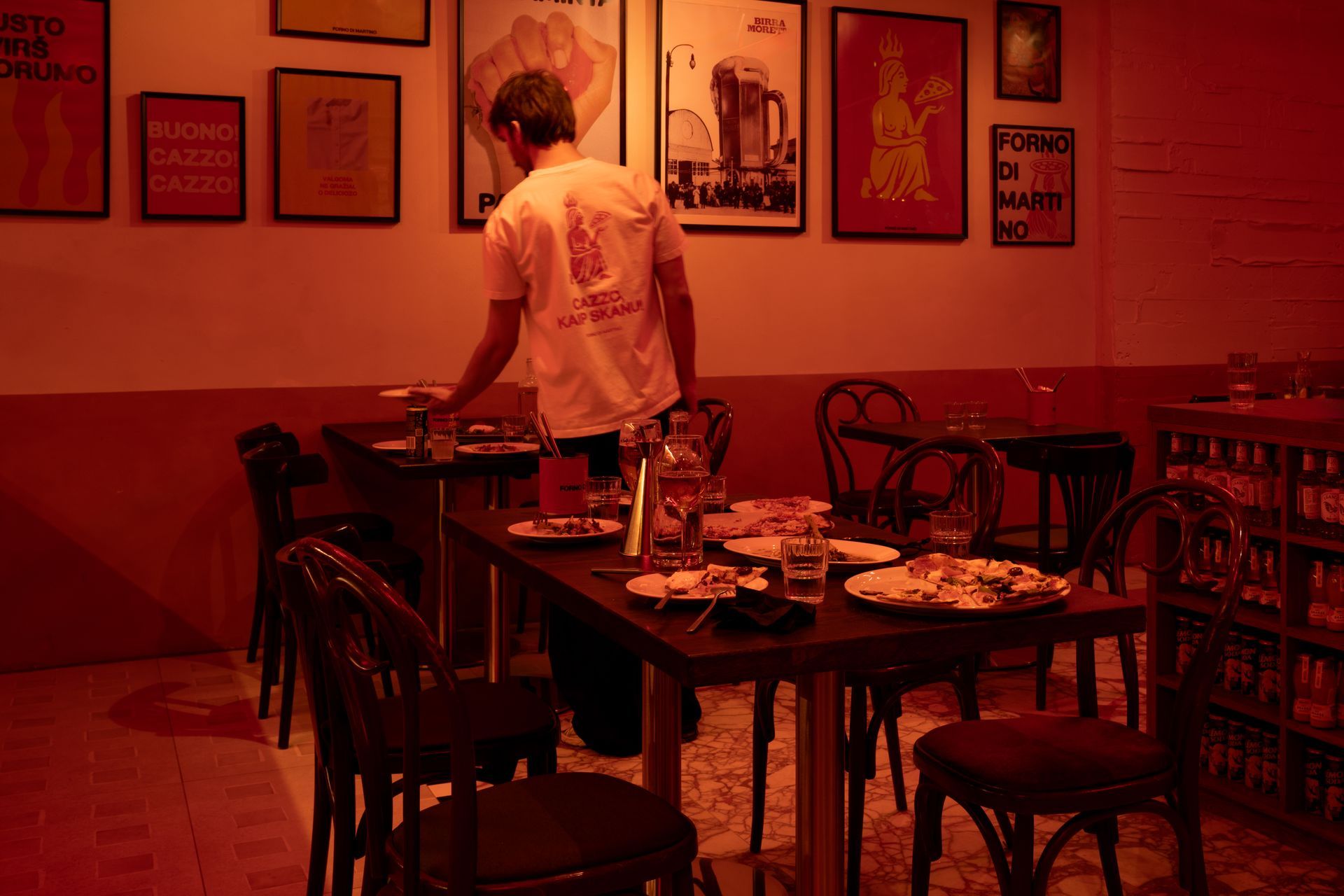 A person in a restaurant, setting a table. The room has posters on the wall and dark furniture.