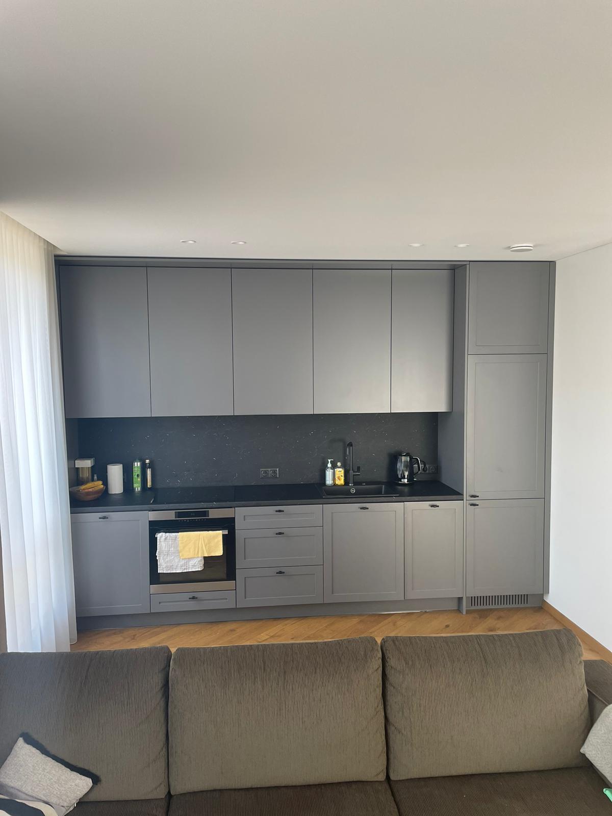 Svetainė su sofa ir virtuvė su pilkomis spintelėmis
